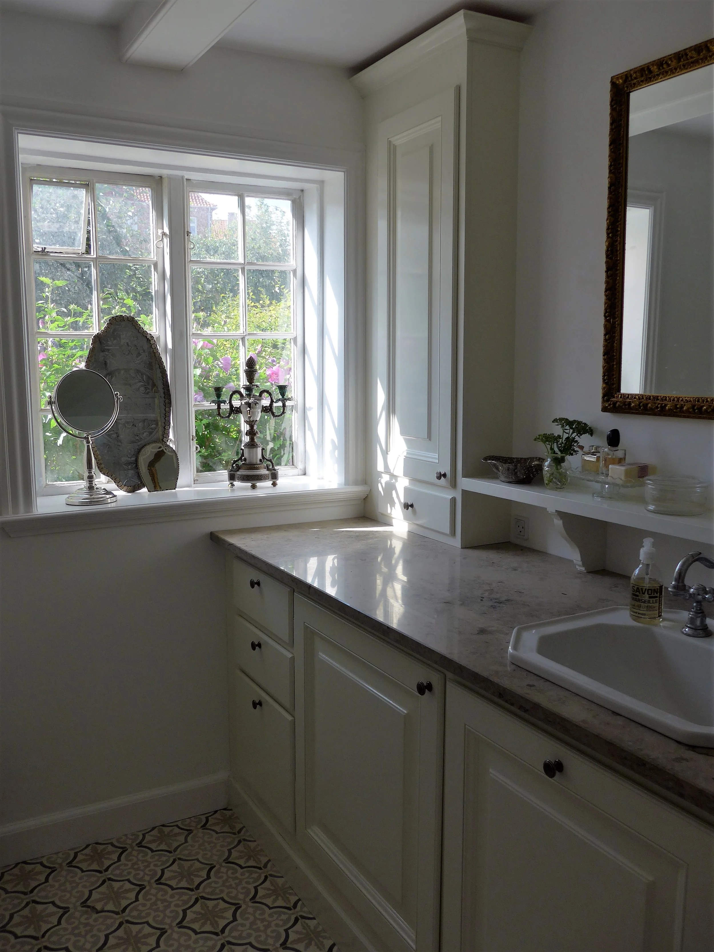 Køkken med hvidt skab, marmorkøkkenbord, vindue med udsigt til blomstrende planter, spejl og toiletartikler på hylde.