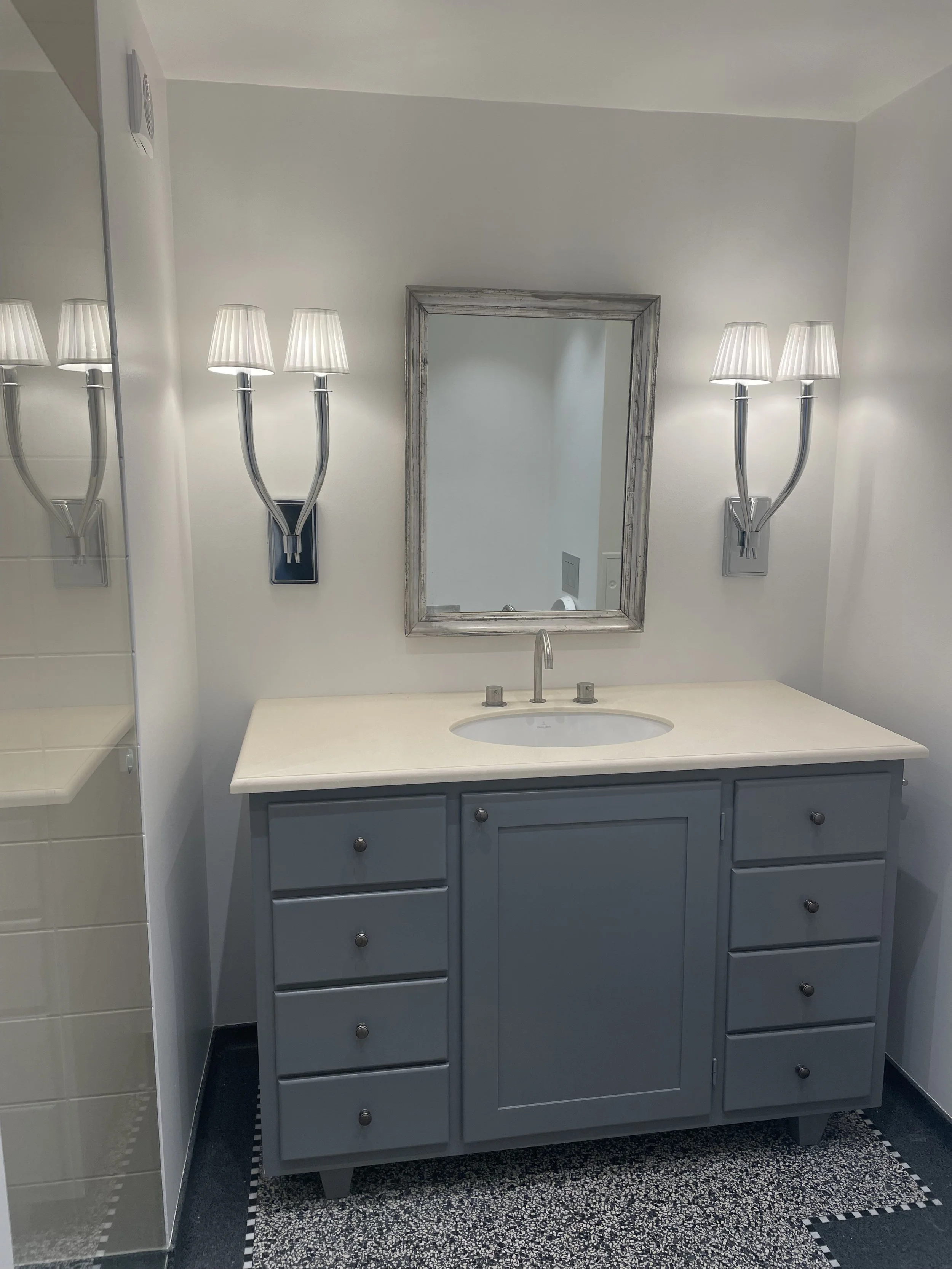 En badeværelsesskab i grå med hvid vask og spejl over, omgivet af væglamper med hvide skærme