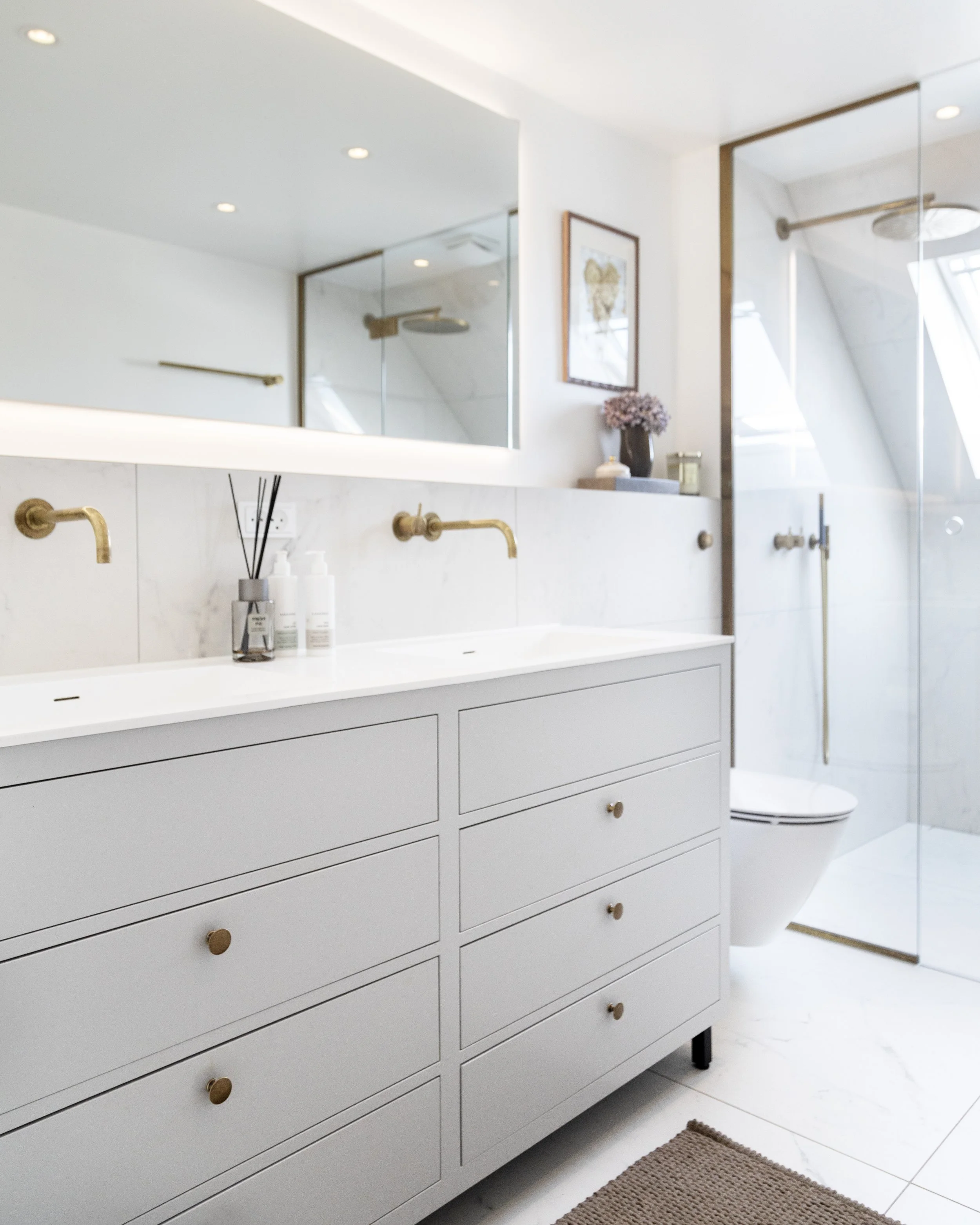 Moderne badeværelse med hvid skabs- og bordplade, guldhaner, stor spejl, brusekabine med glasvæg, toilet, spejllys og dekorationer.