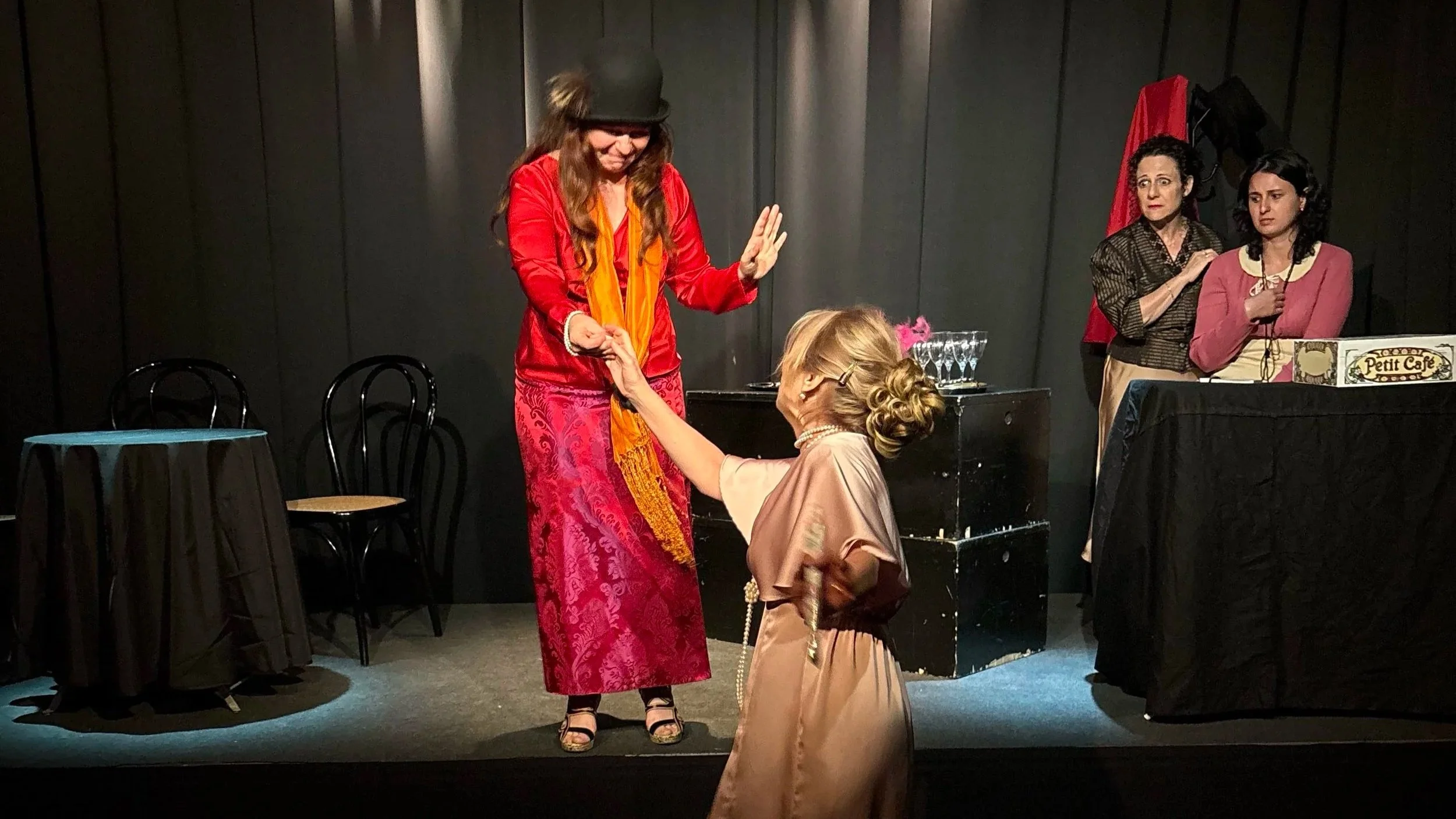 Scene teatrale con tre donne: una in piedi che veste un abito rosso e un cappello nero, due sedute dietro un tavolo con bicchieri. Due donne in piedi osservano con espressioni diverse, una si presenta nervosa e l'altra pensierosa, mentre un'altra donna seduta al centro tiene per mano una donna in piedi.