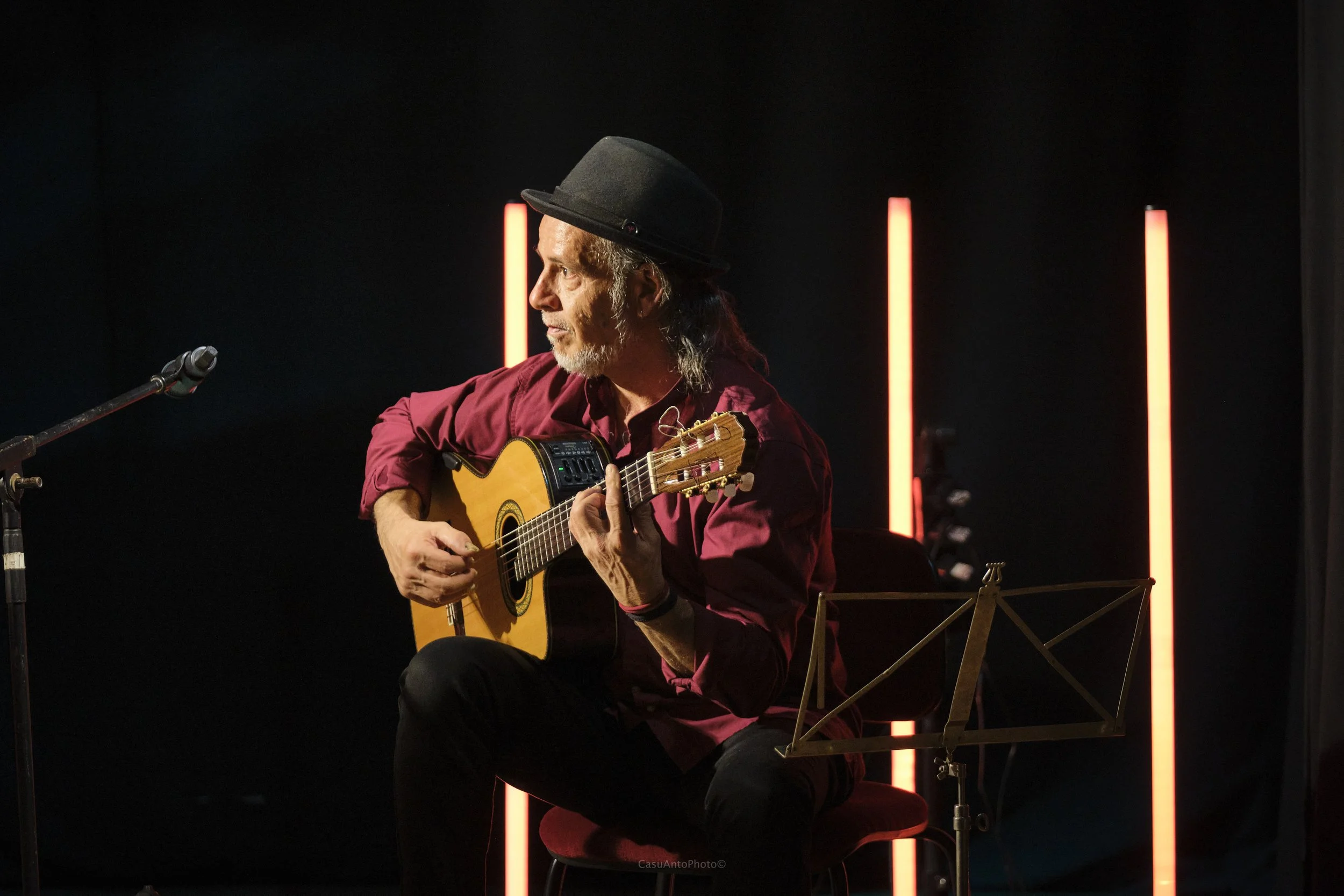 Un uomo anziano con cappello nero suona una chitarra acustica seduto su una sedia rossa, illuminato da luci rosse sullo sfondo in un ambiente scuro.