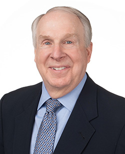 A smiling middle-aged man with light gray hair, wearing a navy suit, light blue shirt, and a patterned blue tie, against a plain white background.
