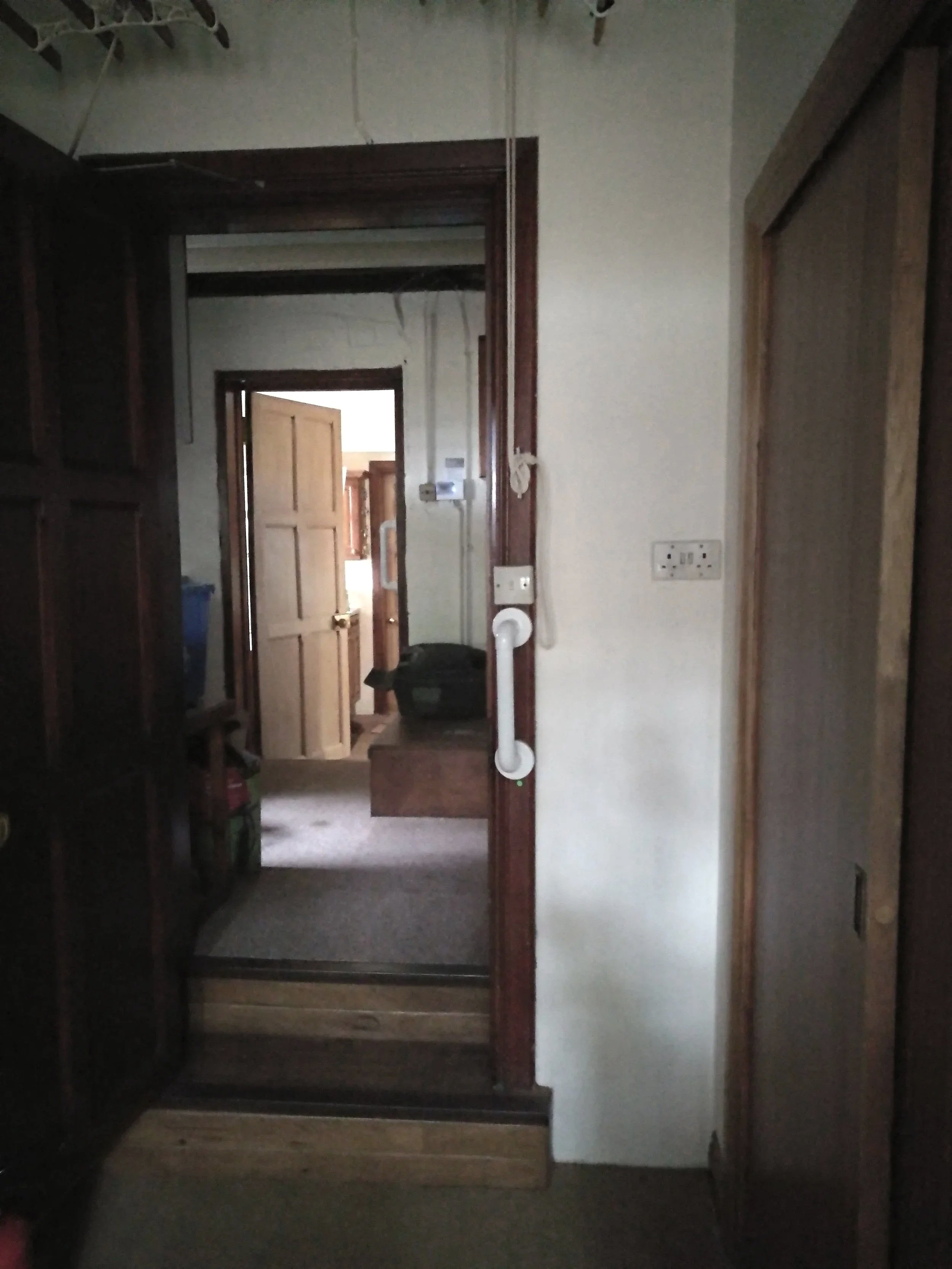 View through a doorway into a small hallway with a staircase, wooden door slightly open at the end, and various household items.