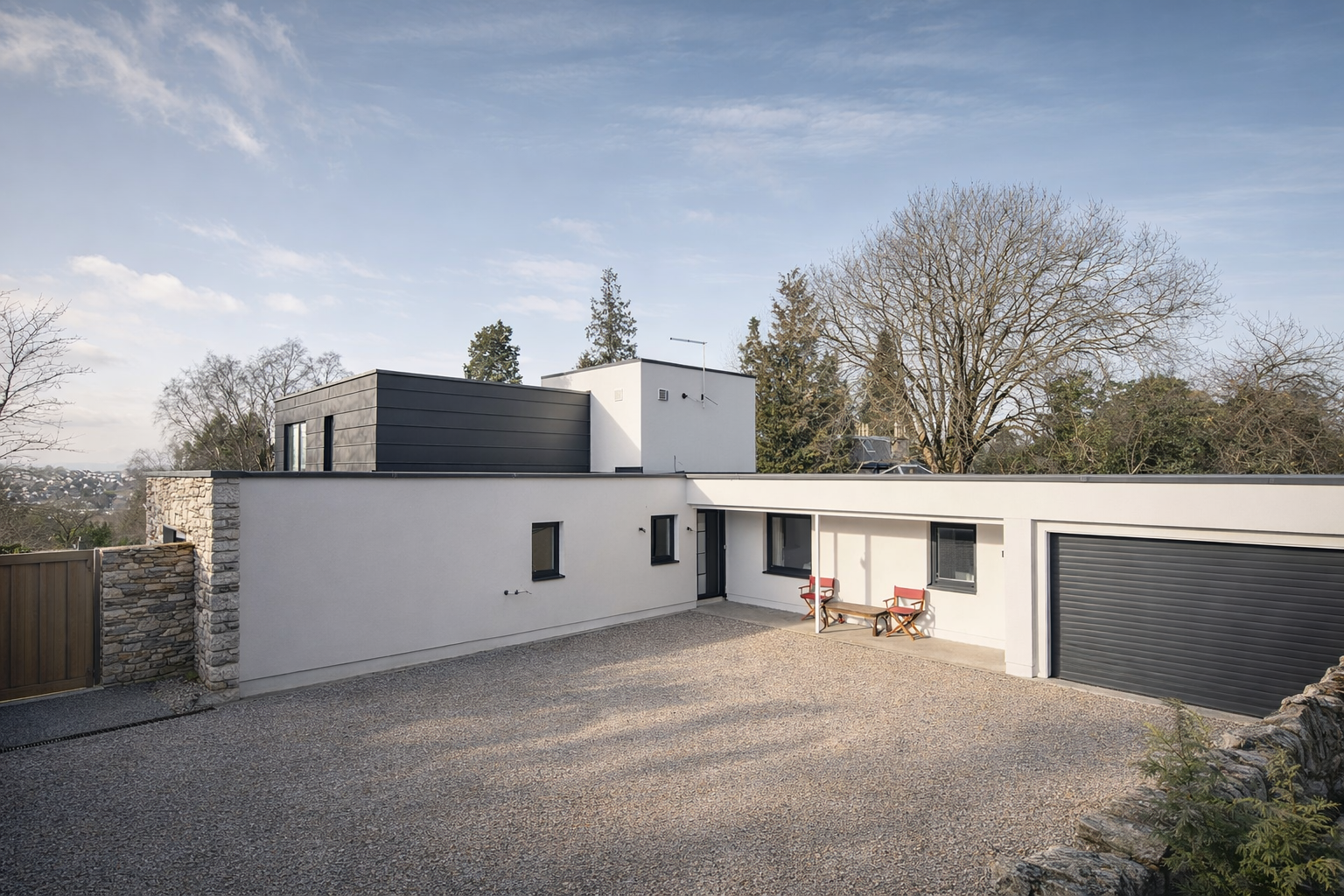 Modern white house with a black garage door, a small patio area with two red chairs and a wooden table, surrounded by trees with leafless branches, under a partly cloudy sky.