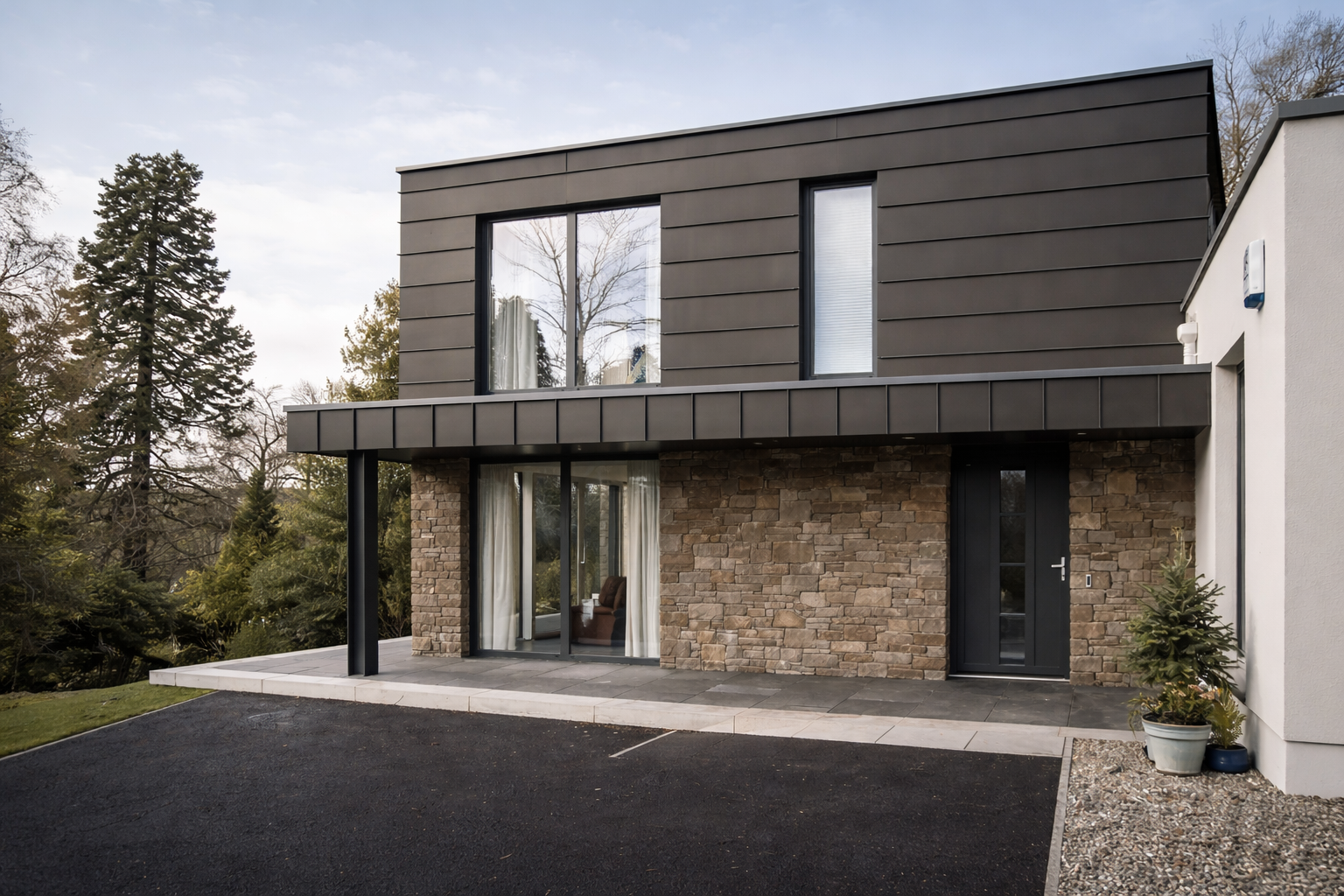 Modern two-story house with black metal siding on the upper level, a brick facade on the lower level, and large windows. There is a small porch area with sliding glass doors, a black door on the right, and a potted plant near the entrance. The driveway is paved and landscaped with grass and gravel.