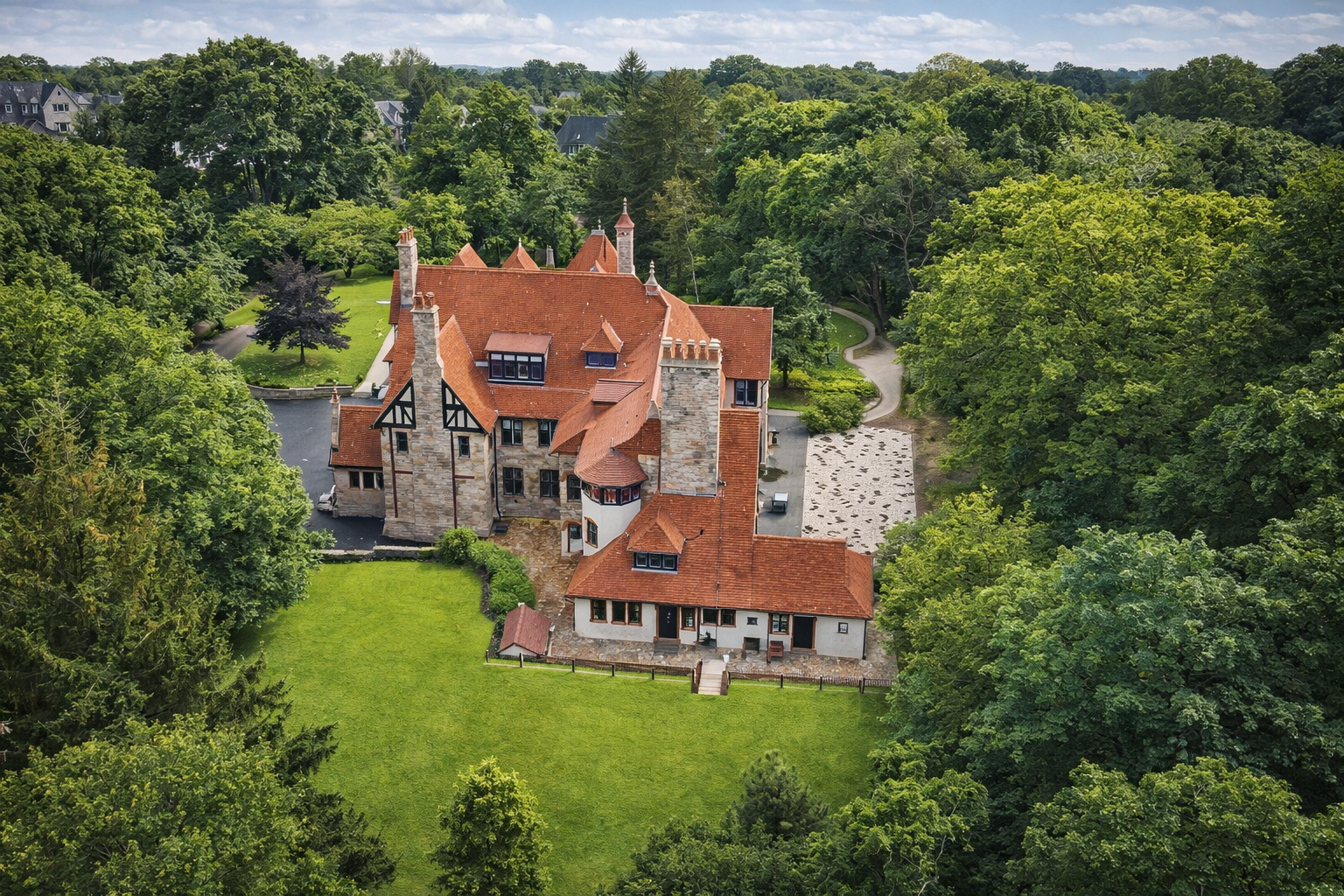 A large, historic house with a red-tiled roof, stone and stucco walls, and multiple chimneys, surrounded by a lush green landscape with tall trees, a well-maintained lawn, and a winding driveway.