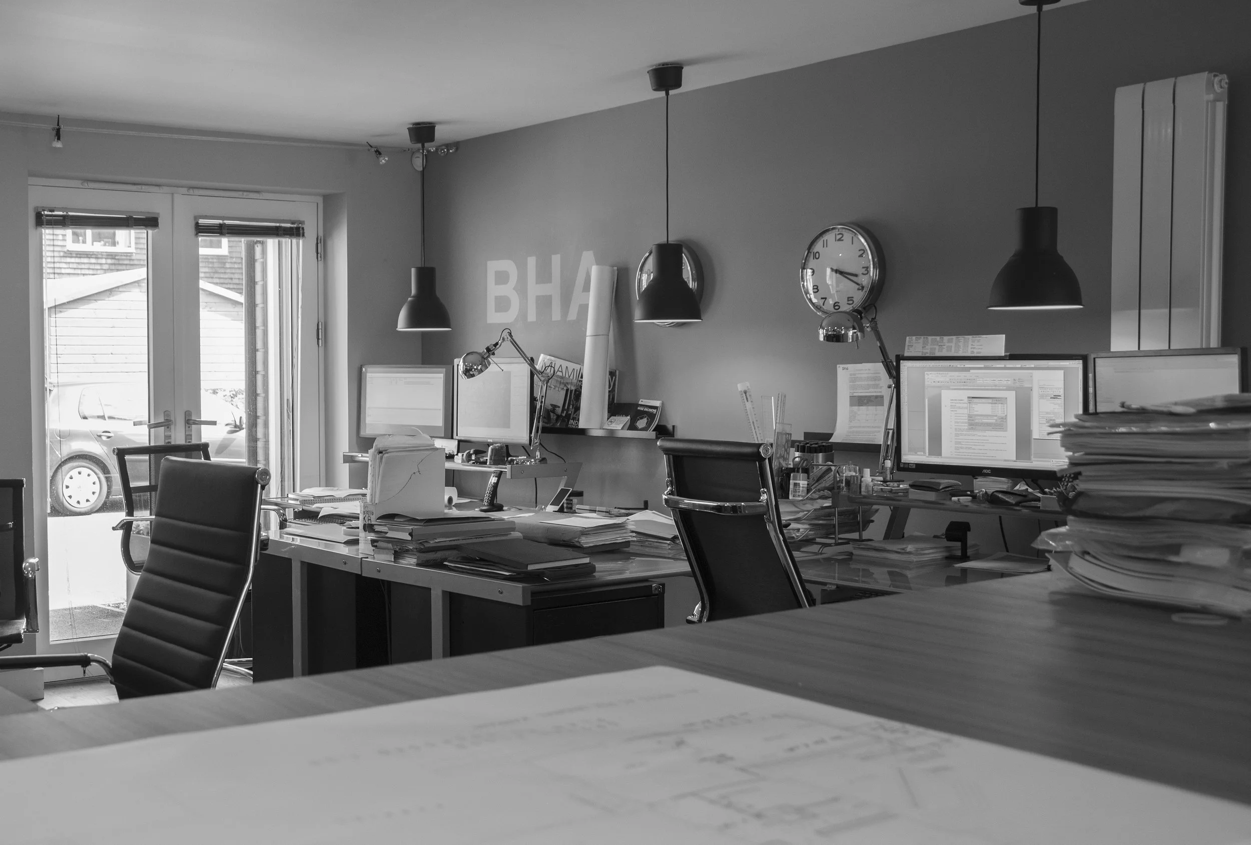 An office space with desks, chairs, multiple computer monitors, stacked papers, and hanging pendant lights. There are French doors leading outside and a wall clock showing 3:14. The room appears organized but busy.