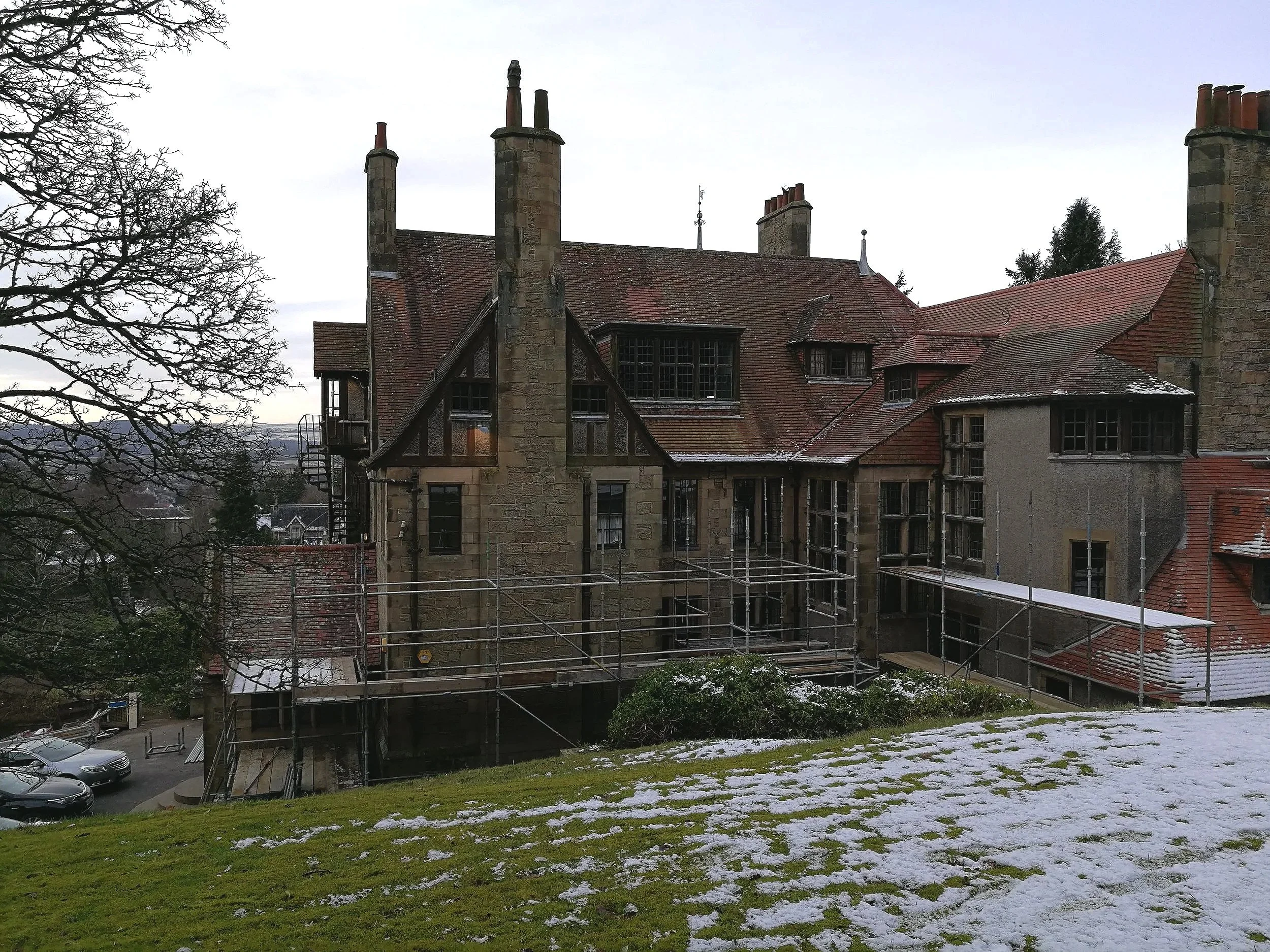 Historic stone building with multiple chimneys, red-tiled roof, and scaffolding outside, surrounded by snowy ground and leafless trees in a winter landscape.