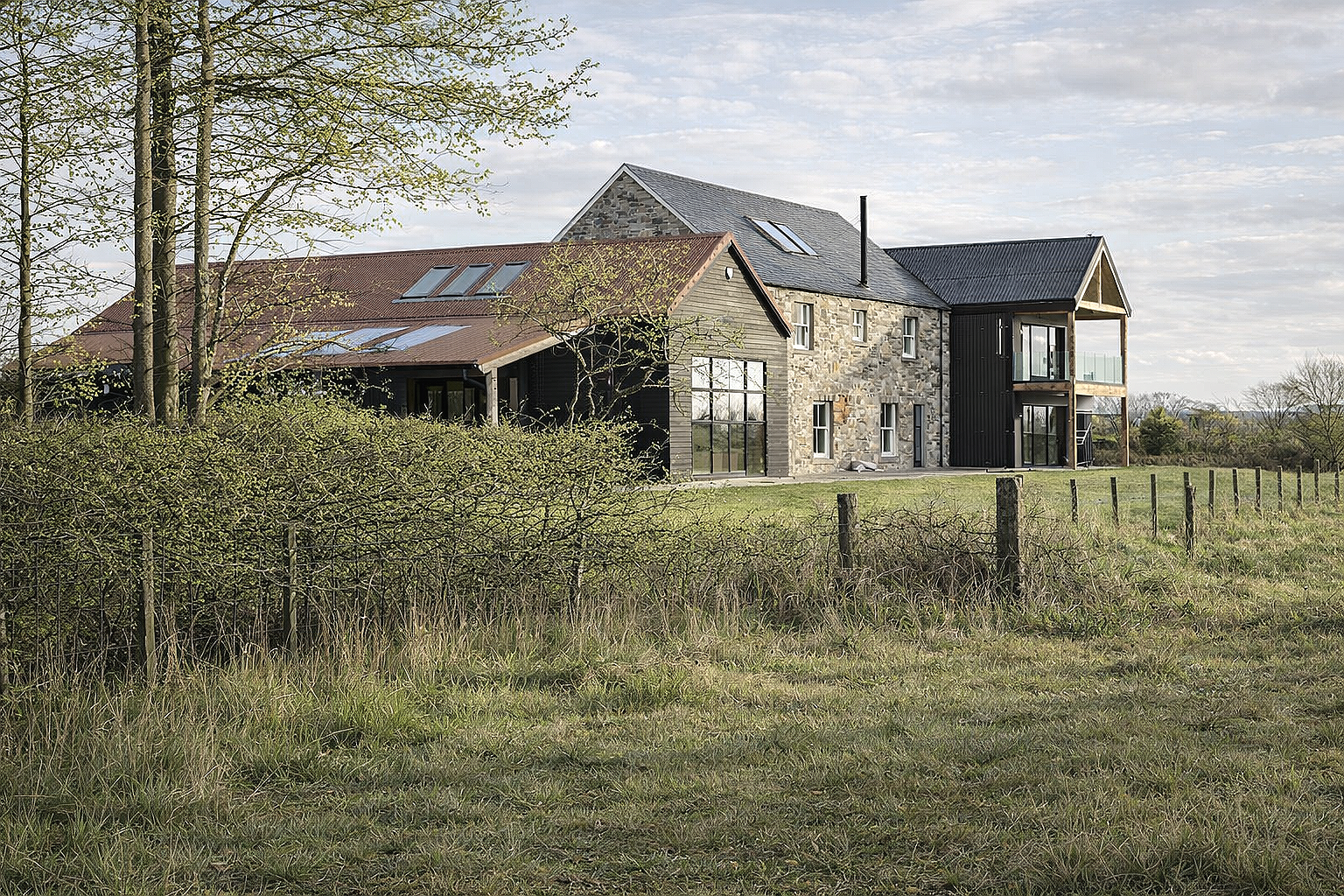 A modern house with a stone and black exterior, large windows, and two balconies, situated in a grassy field with trees and fencing, under a partly cloudy sky.