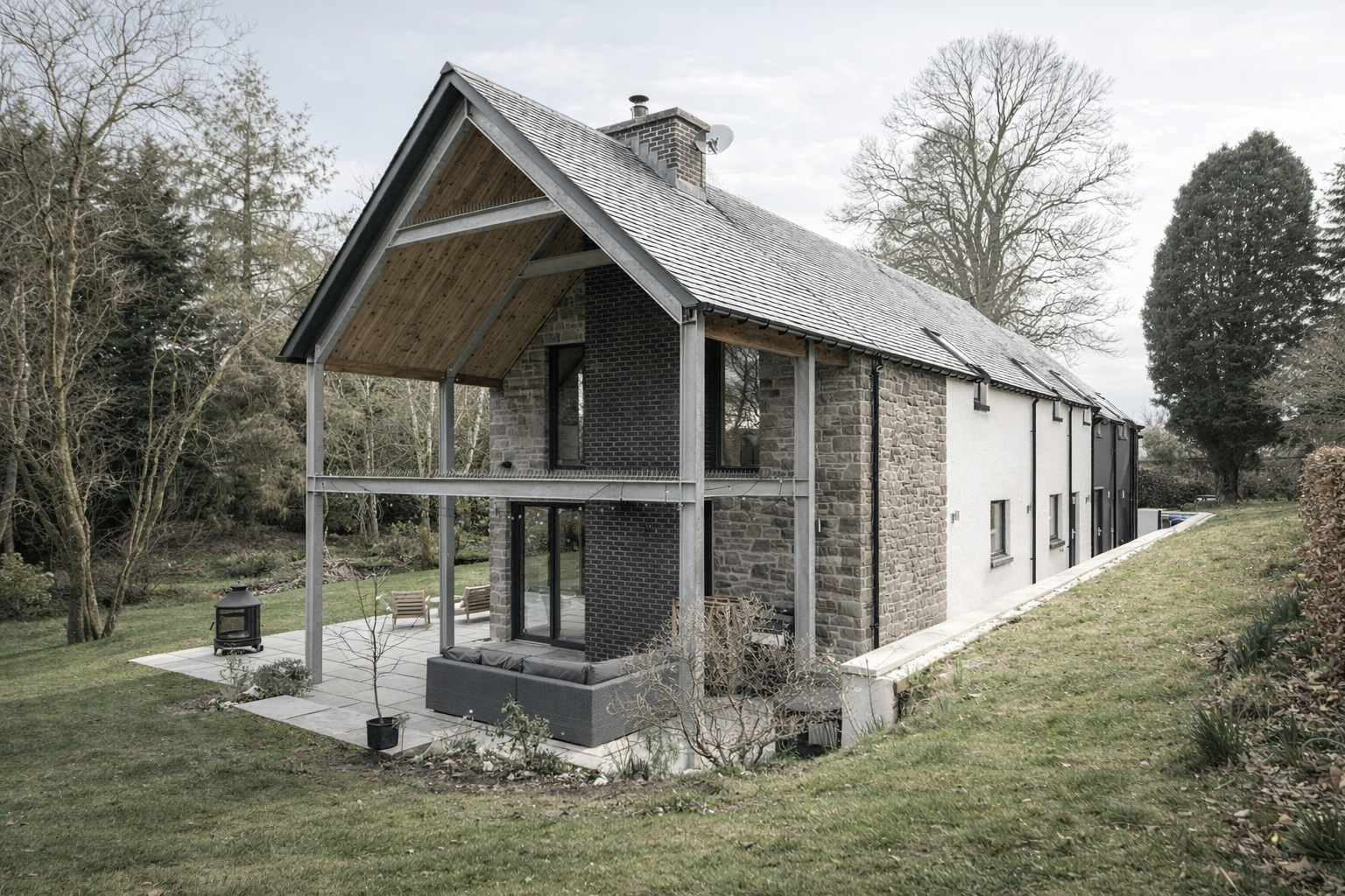 A modern house with gray brick and white stucco exterior, large windows, and a sloped roof with snow, set in a grassy yard with trees and outdoor seating.
