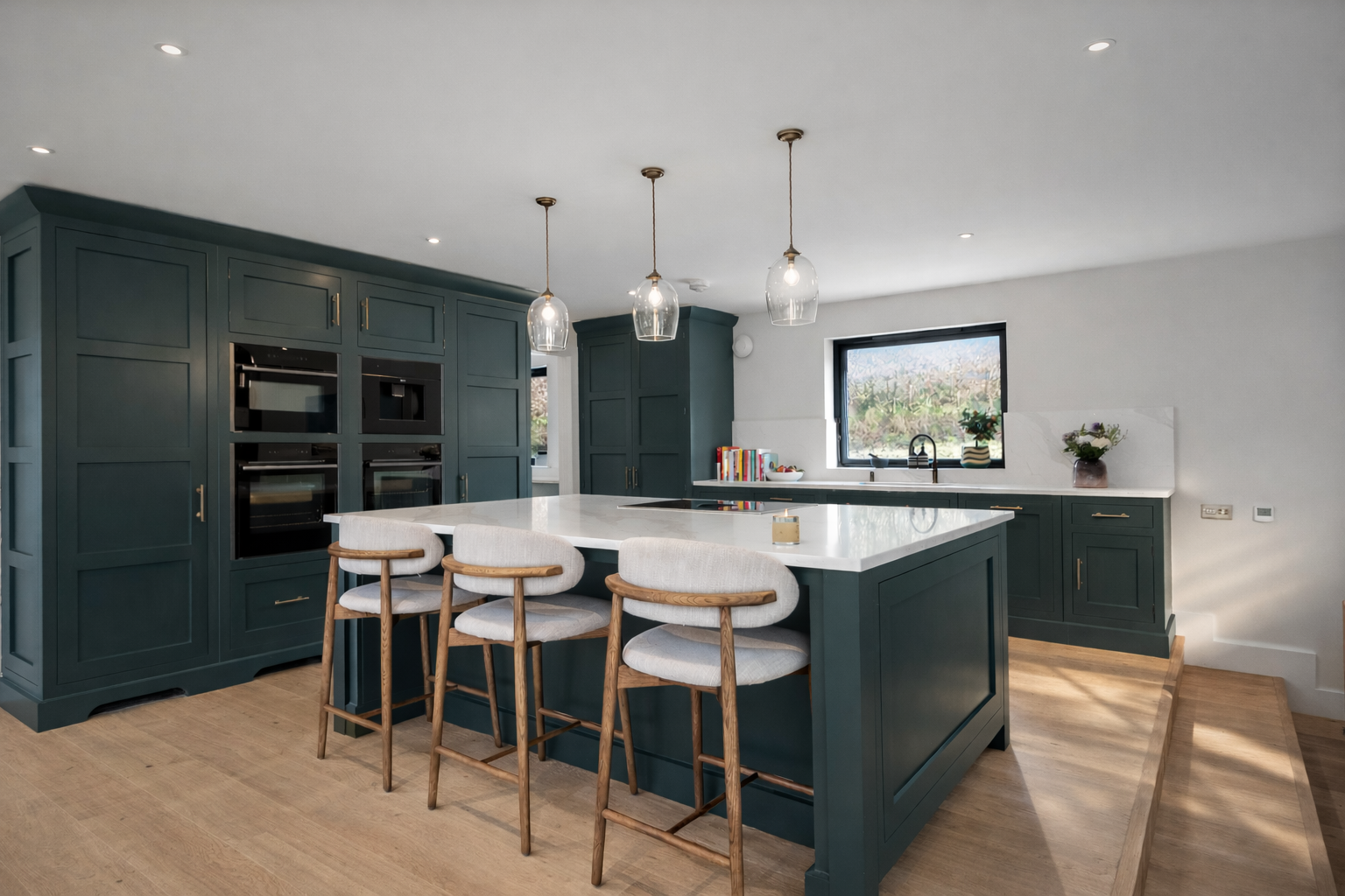 Modern kitchen with teal cabinets, white marble island, four barstools with wooden frames and light upholstery, black-framed window, and wooden flooring.