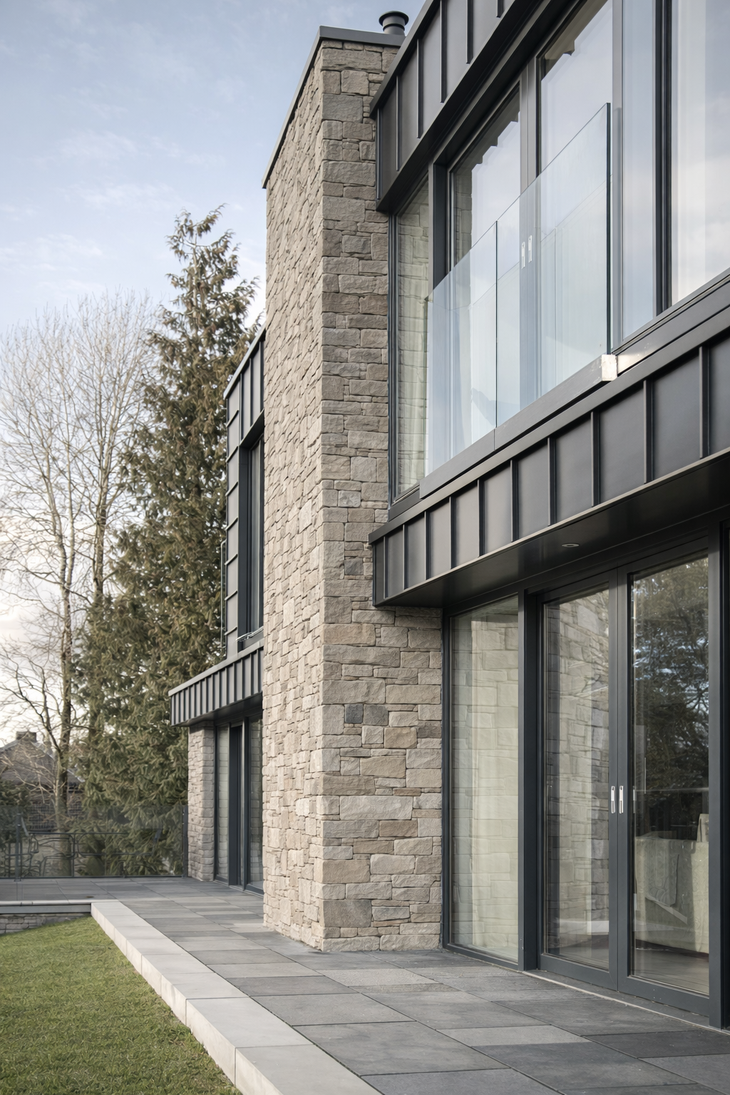Modern house exterior with large glass sliding doors, stone wall, black metal panels, and a patio with tiles, surrounded by trees and a grassy lawn.