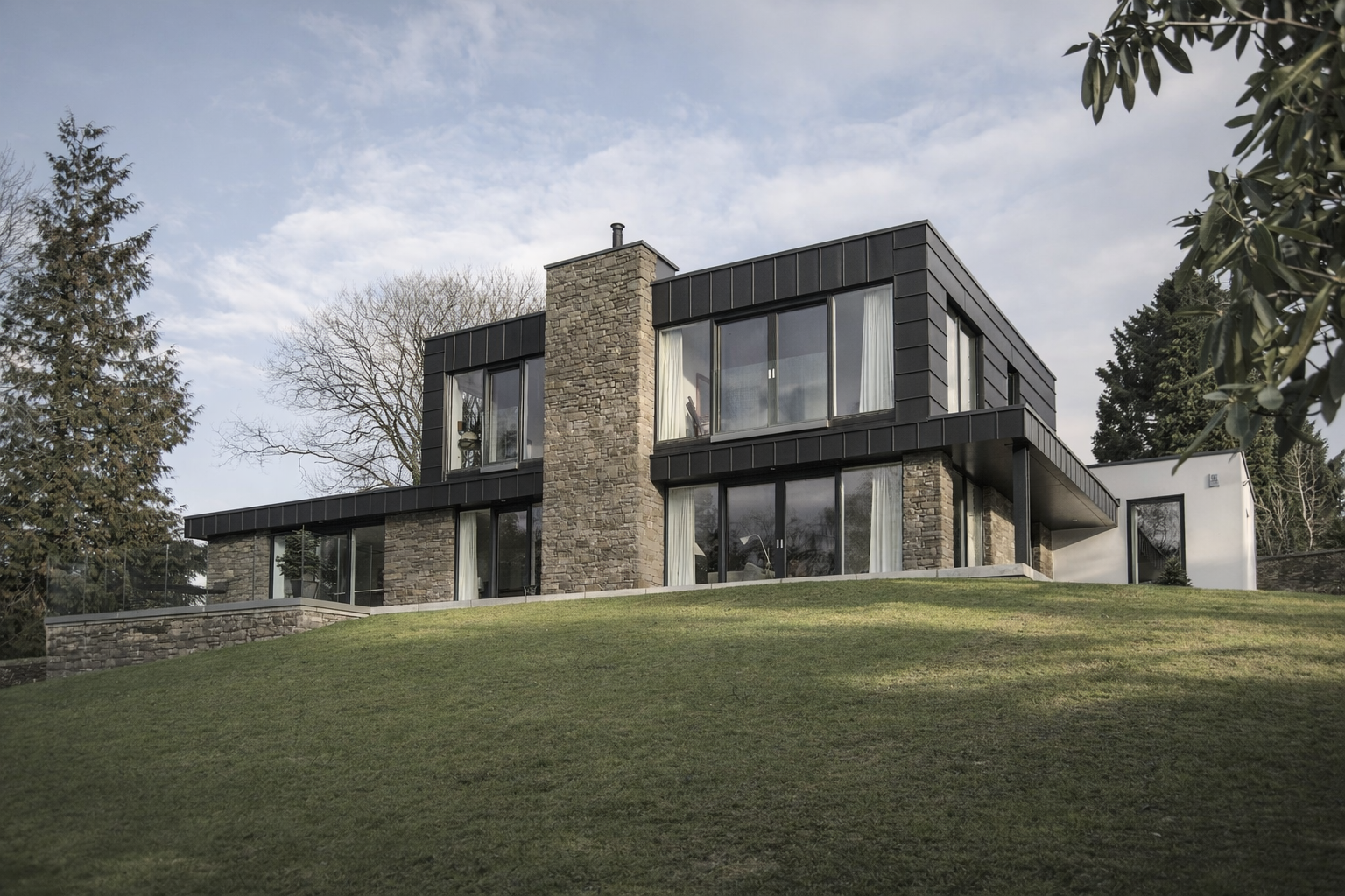 Modern house on a hill with large windows, stone and black exterior, surrounded by trees and a grassy lawn
