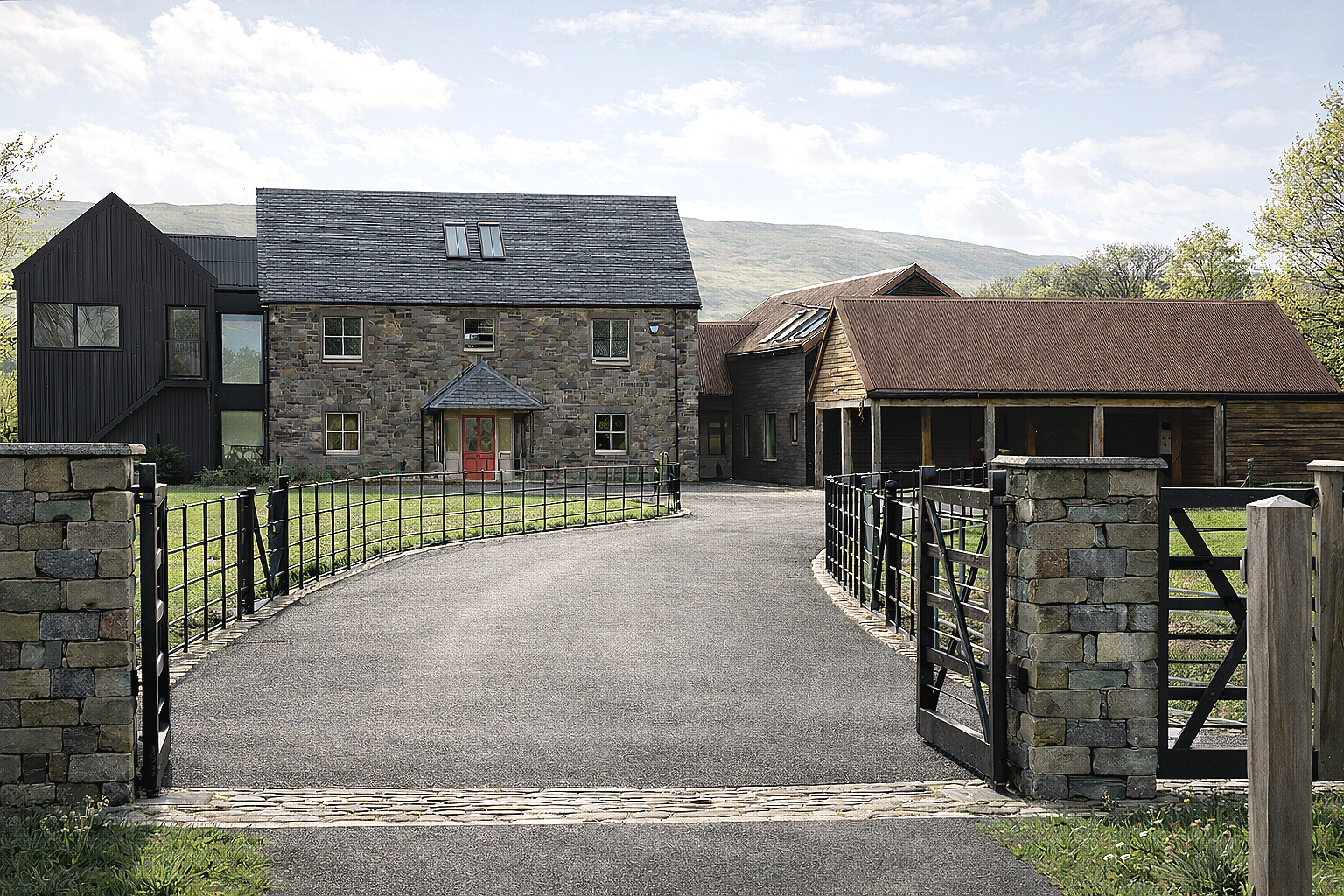 A driveway leading to a house with a stone facade, black siding, and a red front door, surrounded by a stone and metal fence, with a cloudy sky and green hills in the background.