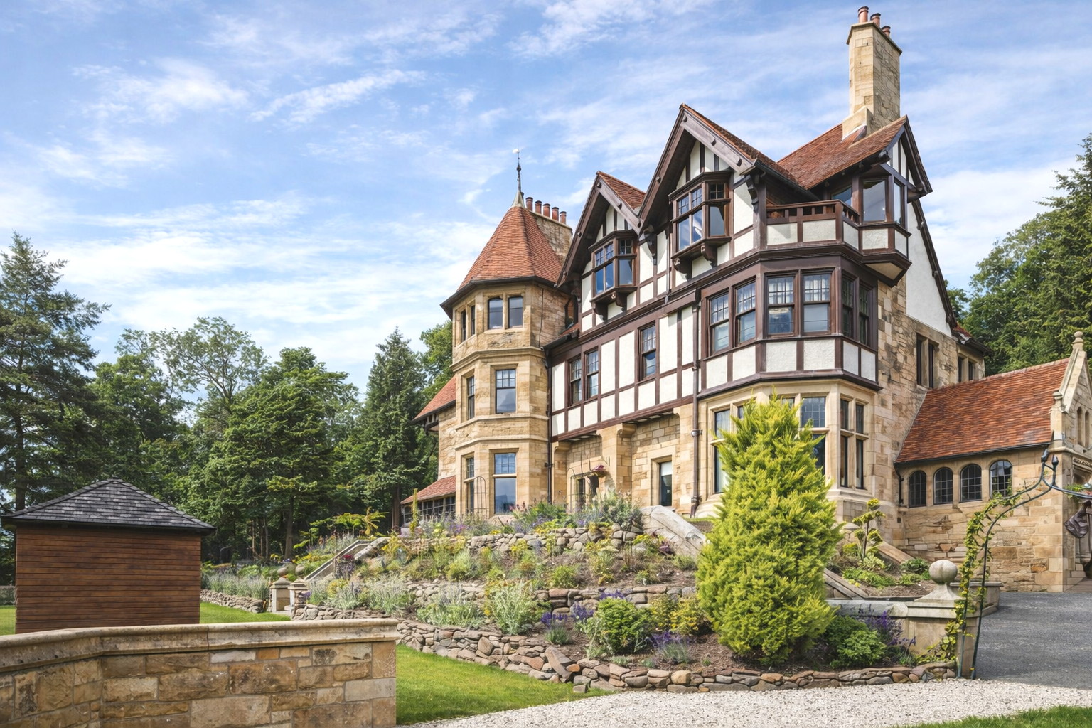 A historic mansion with stone walls, turrets, and a steep red roof, surrounded by landscaped gardens and trees under a blue sky with scattered clouds.