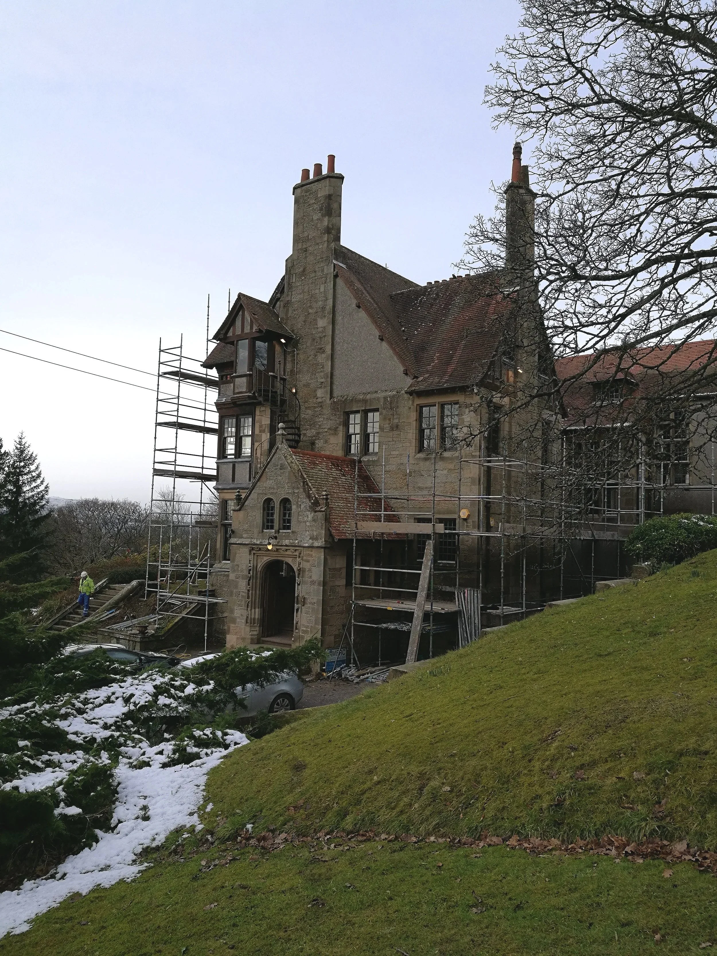 Historic stone mansion undergoing renovation with scaffolding, a car parked nearby, snow on the grass, and a leafless tree in the foreground.