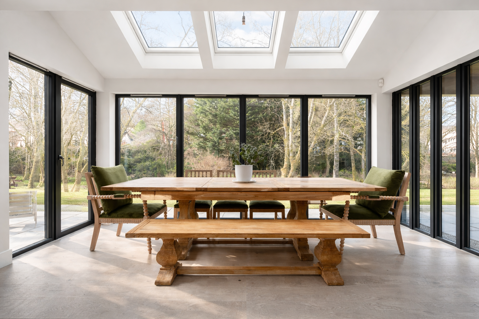 A bright dining room with large window and skylights, featuring a wooden dining table, green cushioned chairs, and a white potted plant in the center, overlooking a lush outdoor garden.