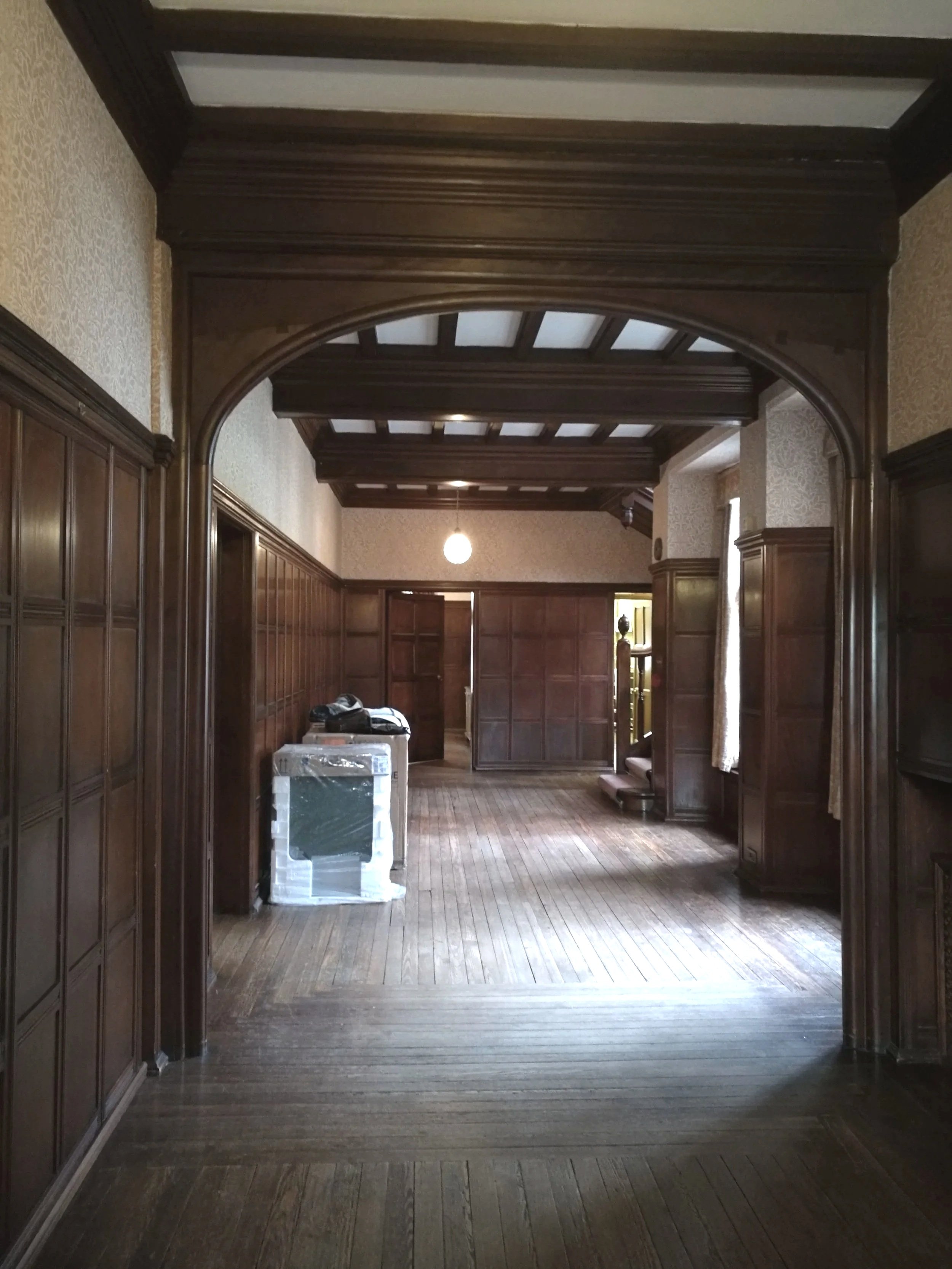 A spacious hallway with dark wood paneled walls, a wooden floor, and a coffered ceiling. There is a round light fixture hanging from the ceiling, and some furniture, including an item wrapped in plastic, is placed to the left.
