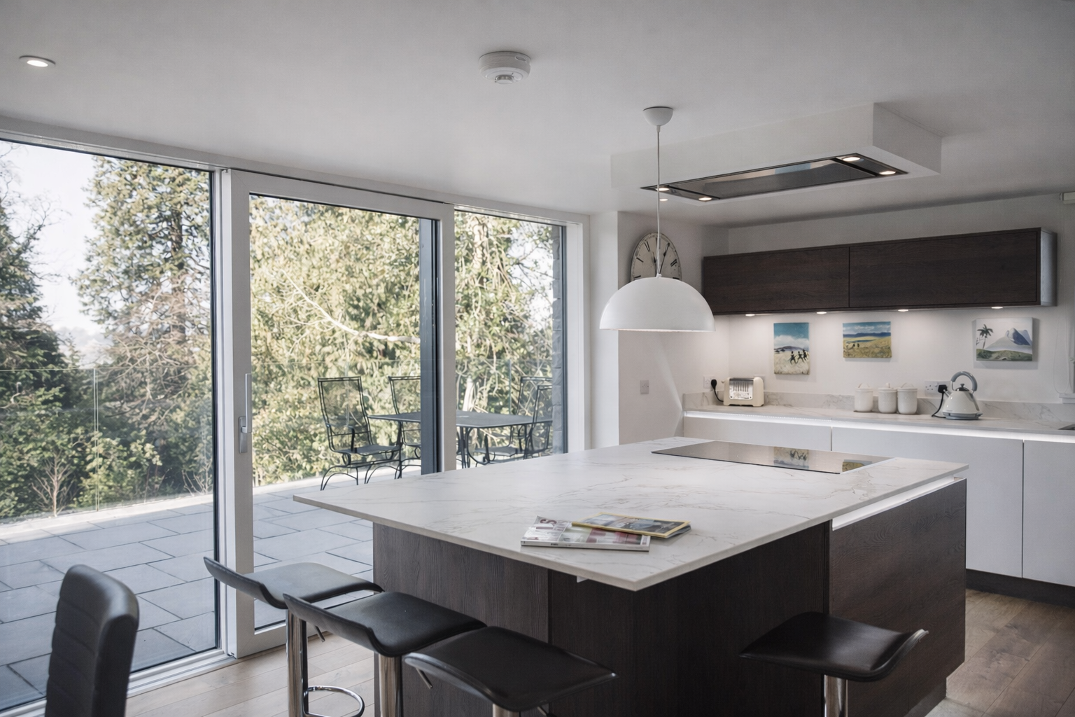 Modern kitchen with white cabinetry, dark wood upper cabinets, and a marble island. Large sliding glass doors lead to a patio with outdoor seating, with trees outside. Kitchen has a hanging white pendant light, small art pieces on the wall, and appliances including a toaster, electric kettle, and a clock.