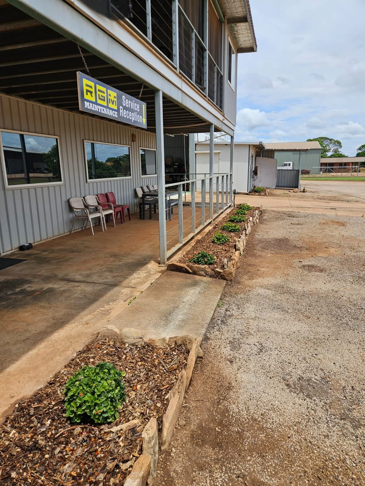 Backyard with palm trees, a chain-link fence, and a small green shed. The yard has a grassy area, gravel path, and mulched plant bed. A house and more greenery are visible in the background.