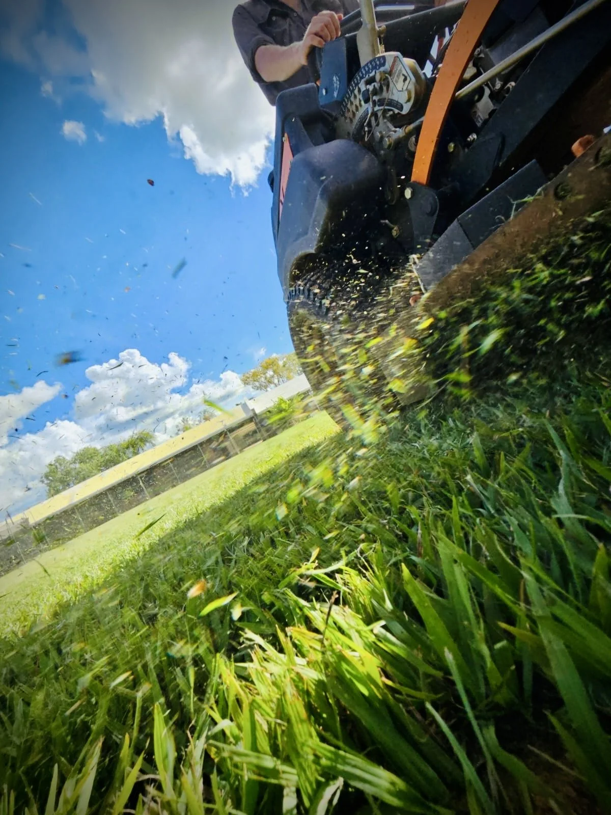 Commercial lawn mower on dirt lot, with trees and buildings in background.