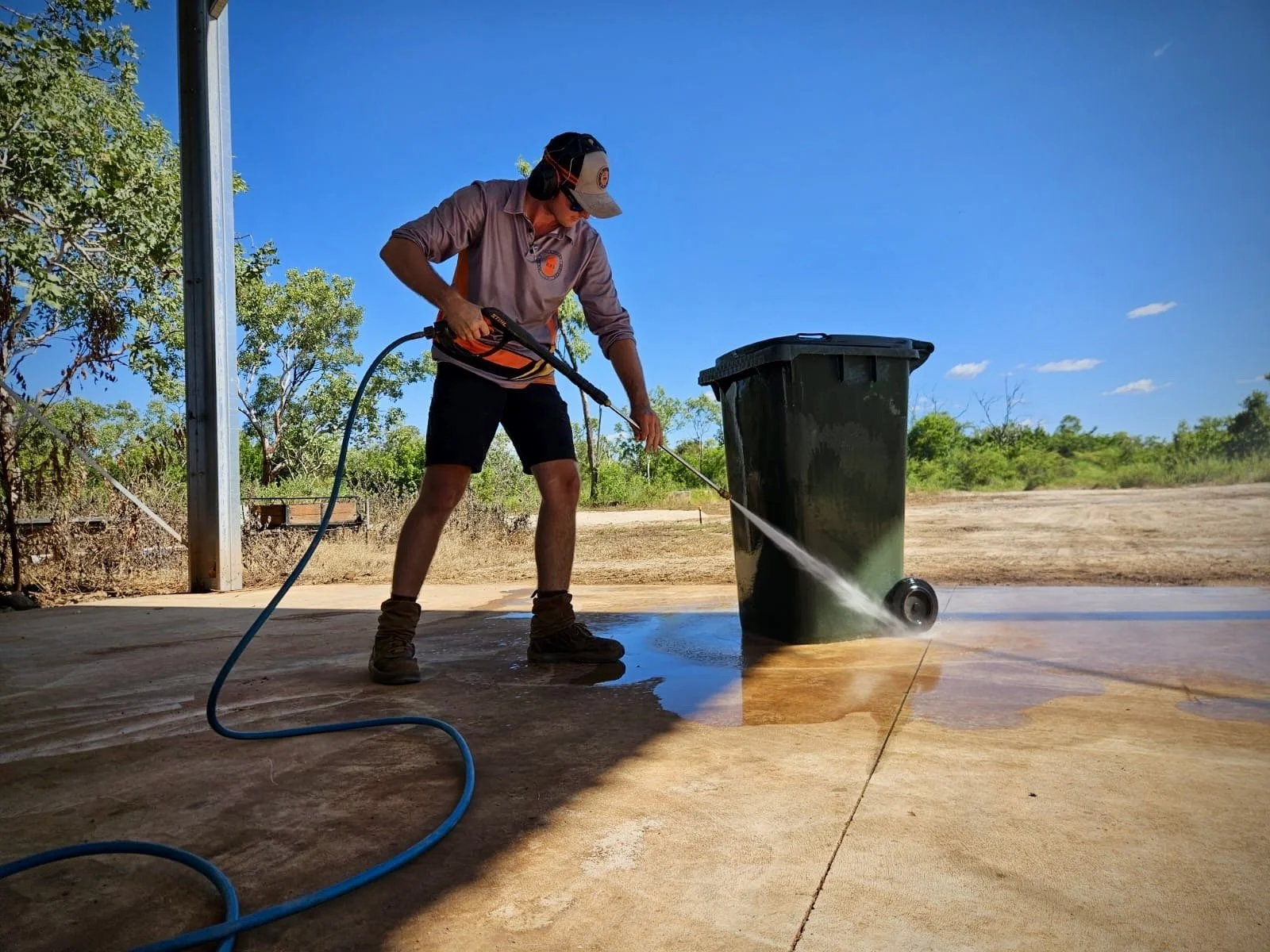 Person using a pressure washer to clean a trash bin outdoors on a sunny day.