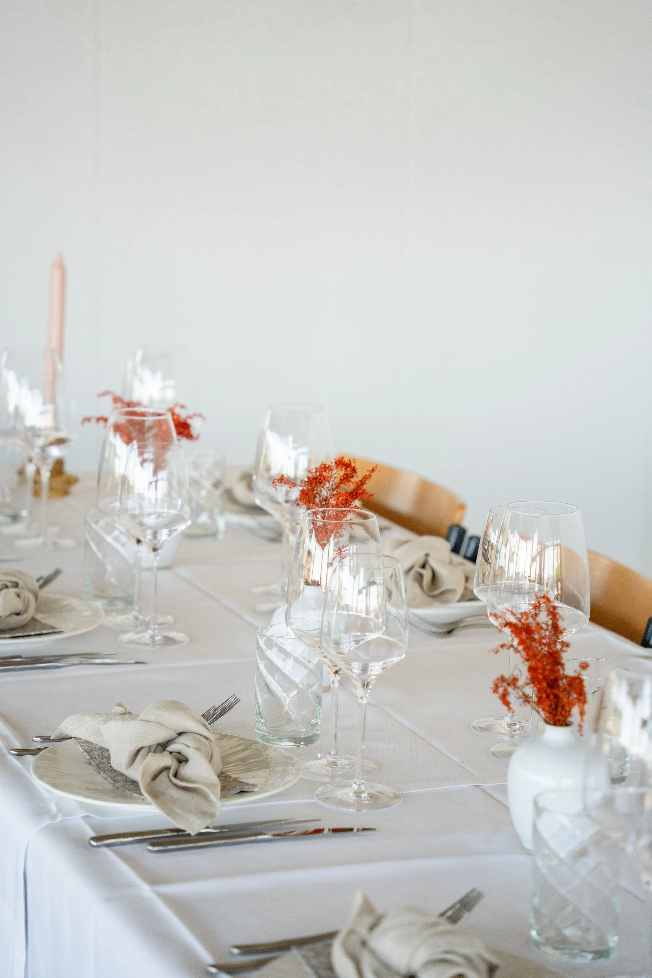 Festbord med hvide duge, tomme vin- og vandglas, beige servietter bundet rundt om bestikket, små vaser med orange blomster, og en lys rosa lydstang i baggrunden.