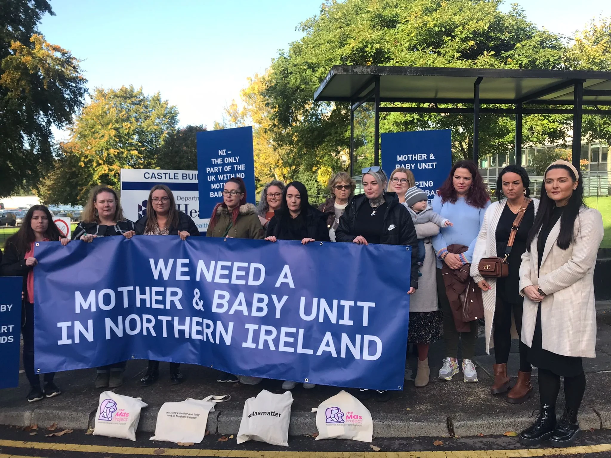 A group of women and babies holding a blue sign with white lettering saying "We need a mother and Baby Unit in NI".
