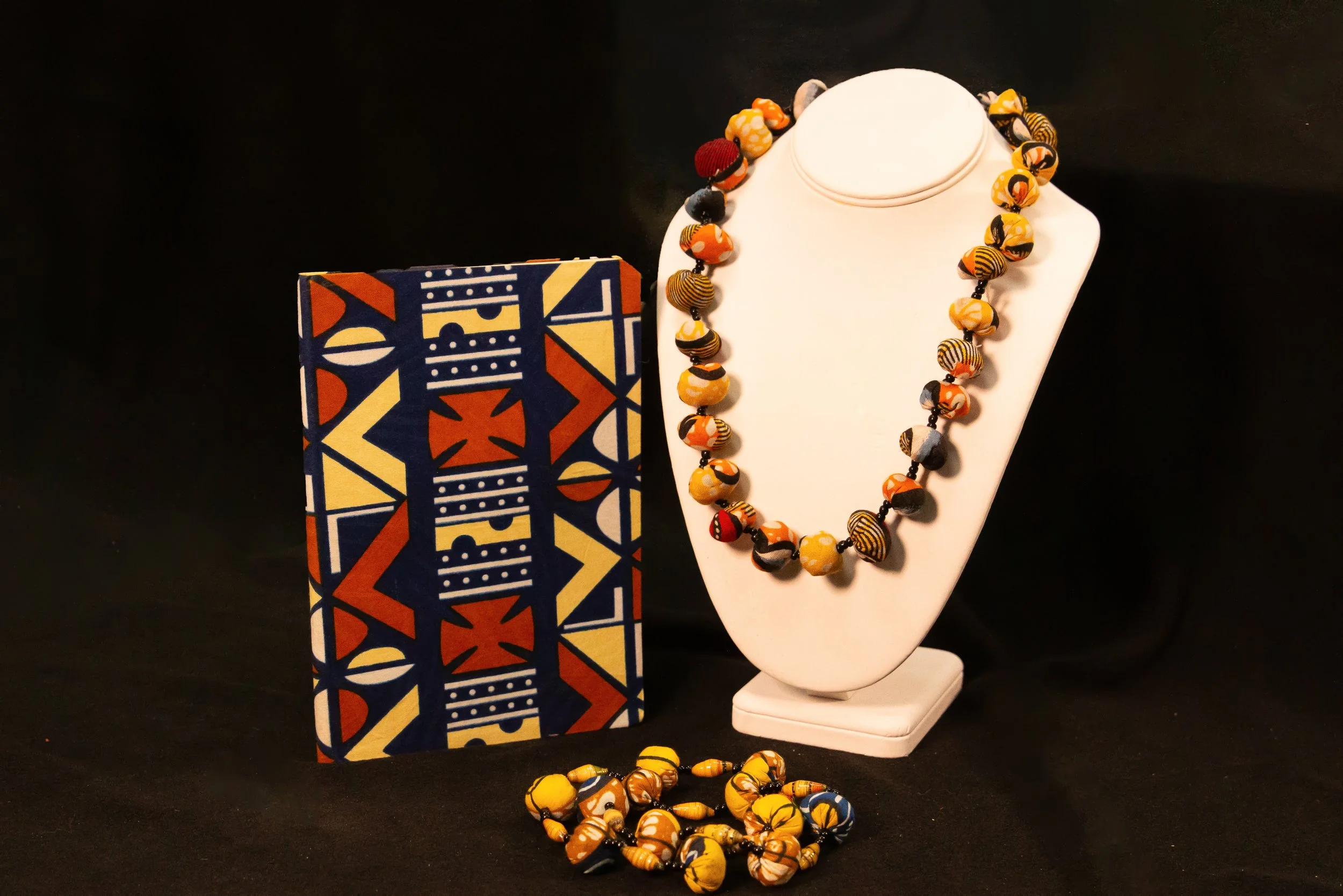 Colorful African-style beaded jewelry set, including a necklace and a bracelet, displayed on a white jewelry stand next to a matching patterned box and loose beads on a black background.