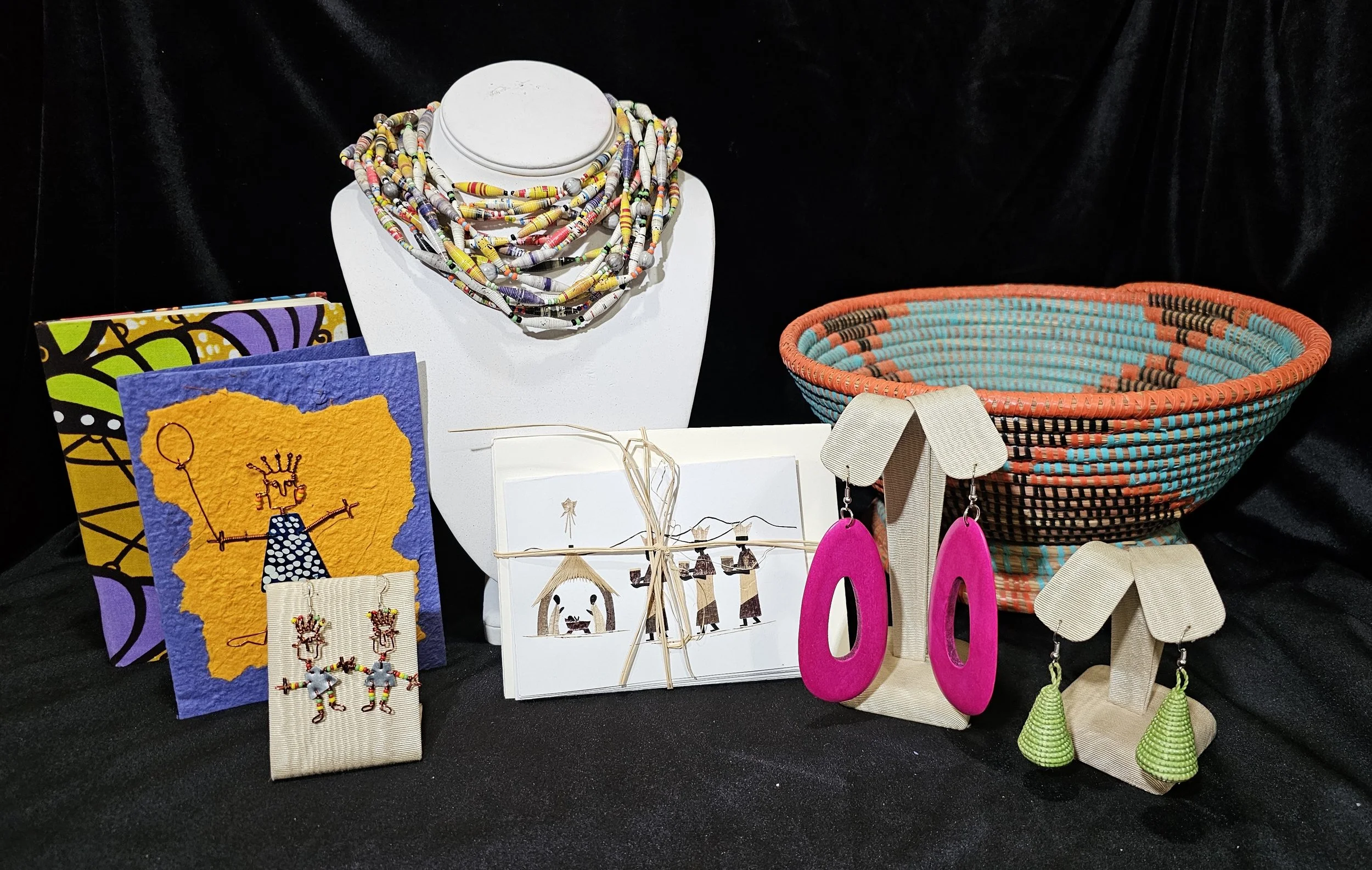 Collection of handmade jewelry, artwork, and crafts displayed on a black surface against a black background. Includes beaded necklaces, earrings, woven bowls, and greeting cards.