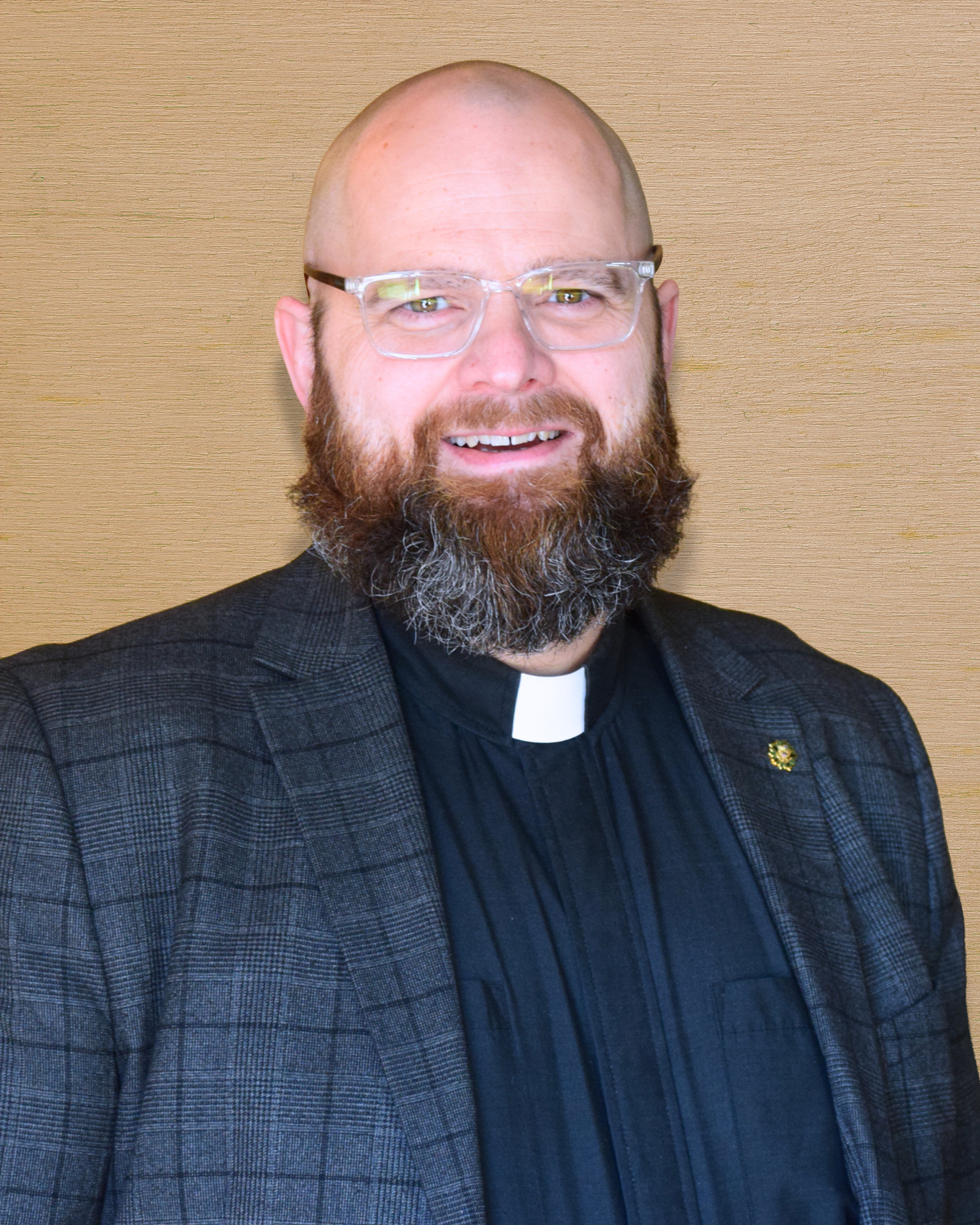 A man with a beard and glasses wearing religious clerical attire and a checked blazer, standing against a wooden textured background.