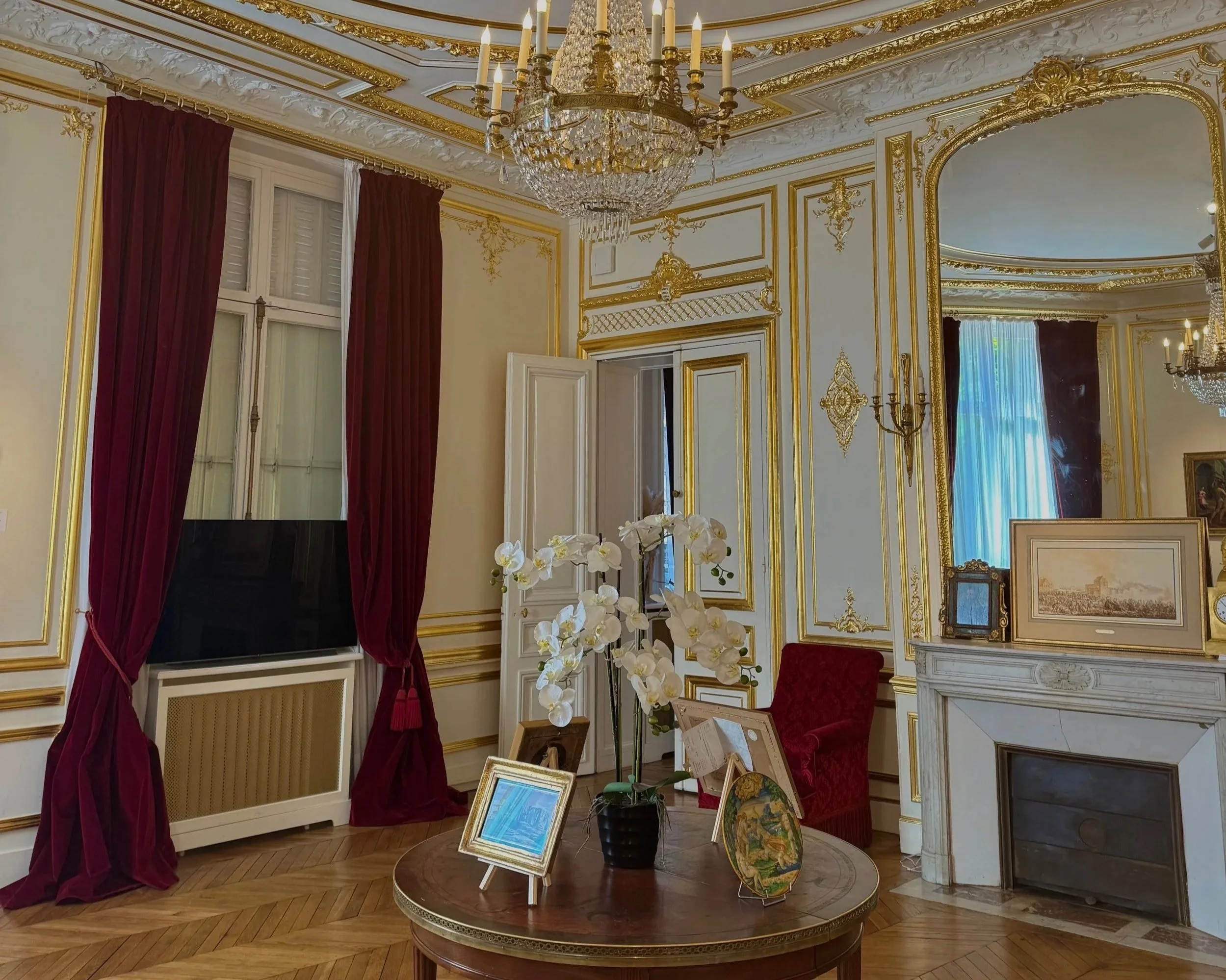 Une salle ornée avec des murs décorés de moulures dorées, un chandelier en cristal suspendu au plafond, une cheminée, une table ronde avec des cadres et une orchidée, des rideaux rouges et des fenêtres avec volets. Un miroir et un fauteuil en velours rouge sont également visibles.
