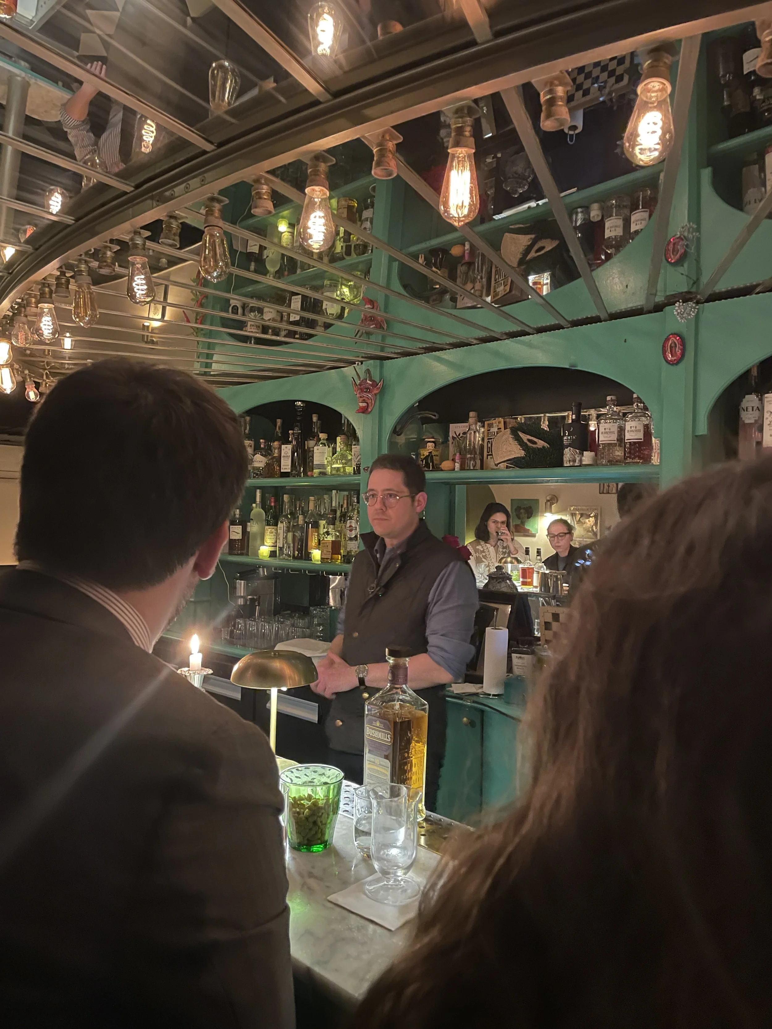 Une scène dans un bar avec un barman derrière le comptoir, des bouteilles d'alcool sur des étagères, des clients en première ligne, un éclairage tamisé avec des ampoules suspendues, et un décor coloré avec une tête de diable rouge sur le mur.