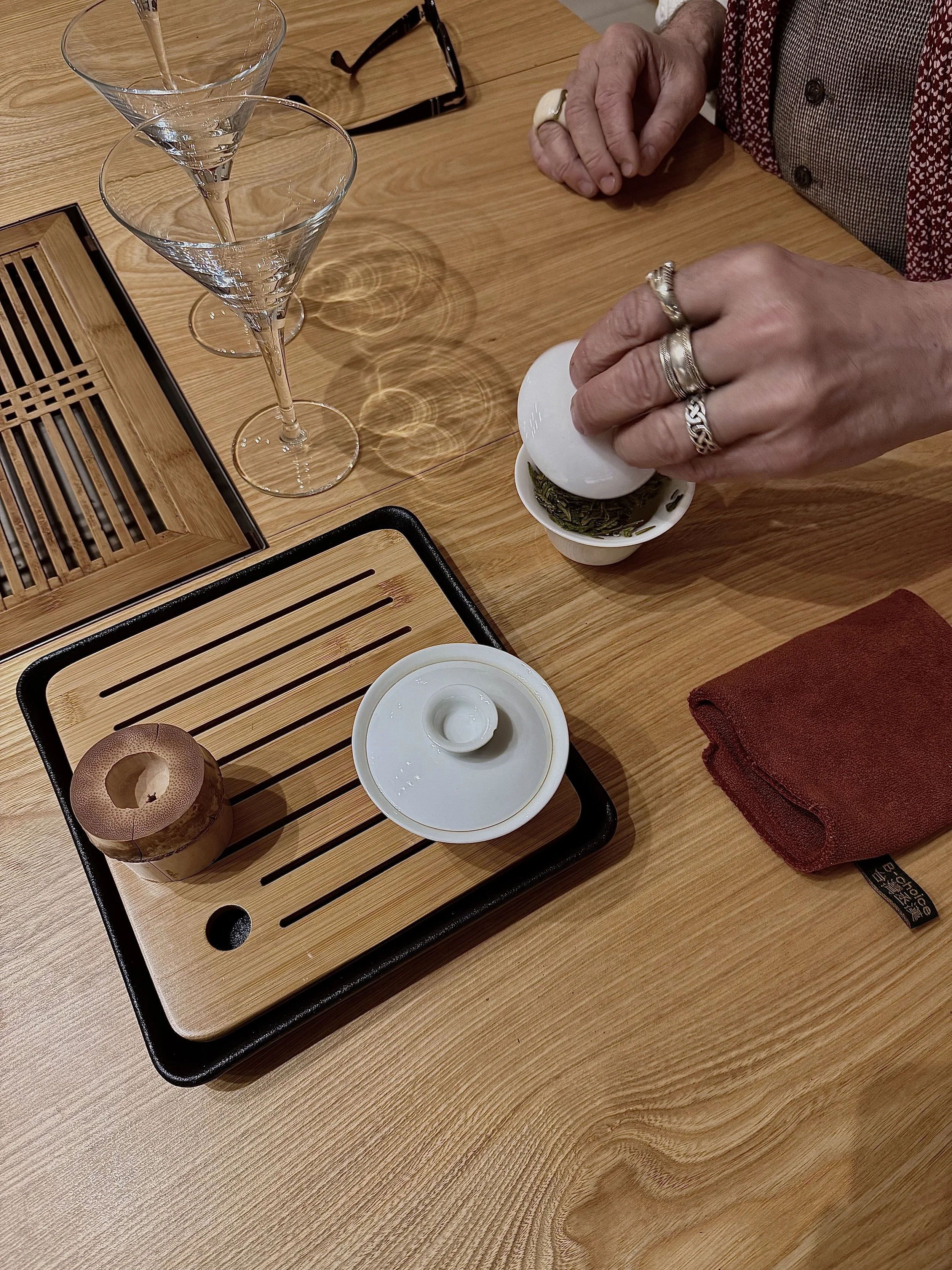 Une personne verse du thé dans une tasse en céramique verte. Sur la table en bois, il y a deux grands verres à cocktail vides, un porte-couteaux en bois, une petite boîte en céramique avec un couvercle, une serviette rouge et une paire de lunettes de soleil. Un tapis en bois est placé sous la vaisselle.