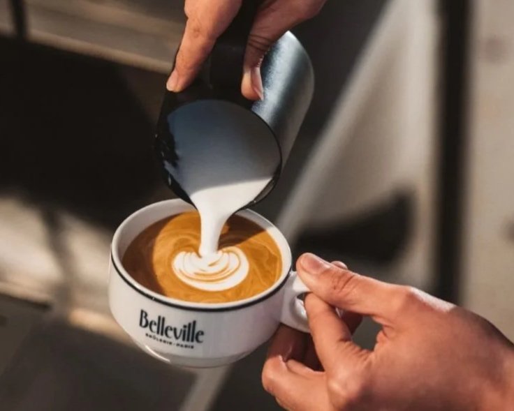 Une main verse du lait chaud dans un mug de café avec du latte art, mug marqué de 'Belleville'.
