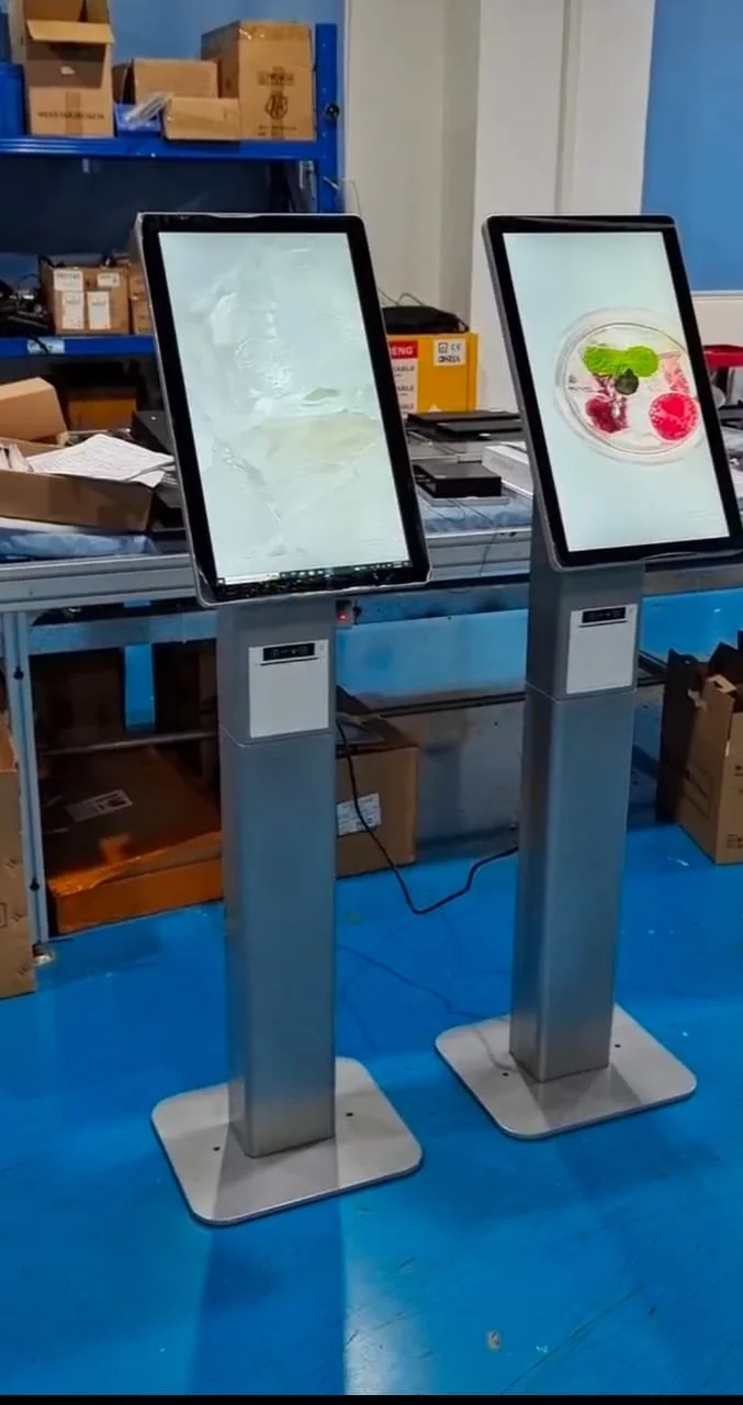 Two digital kiosk screens on stands in a warehouse or factory setting, with boxes and shelves in the background.