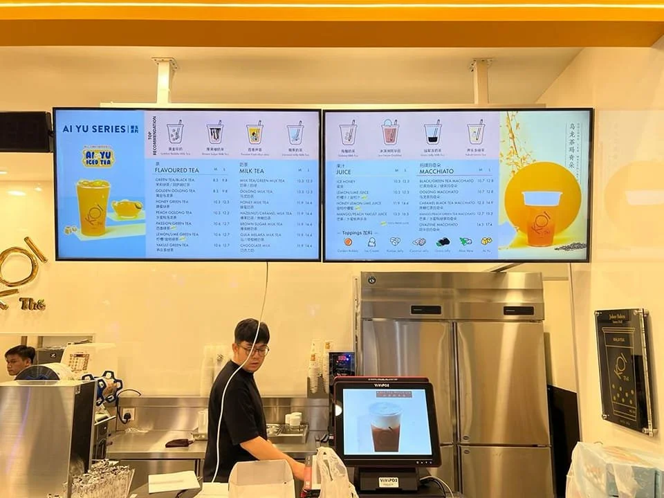 Digital menu boards displaying various tea and juice options at a bubble tea shop. A staff member preparing drinks behind the counter.