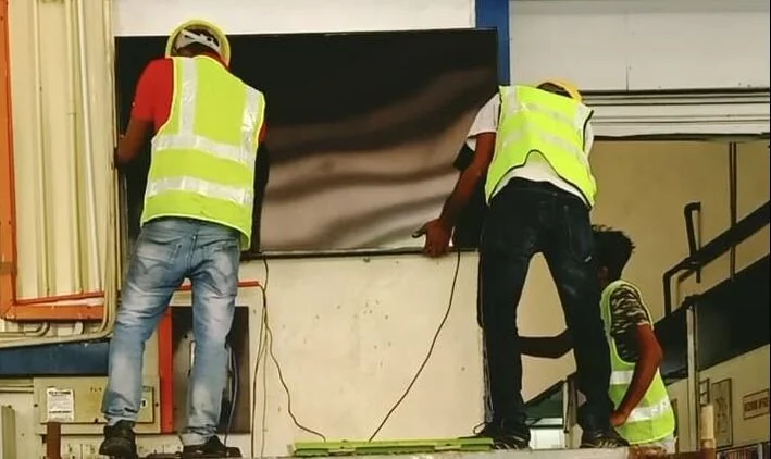 Three workers wearing yellow safety vests and helmets installing or working on a large screen or monitor on a wall inside a building.