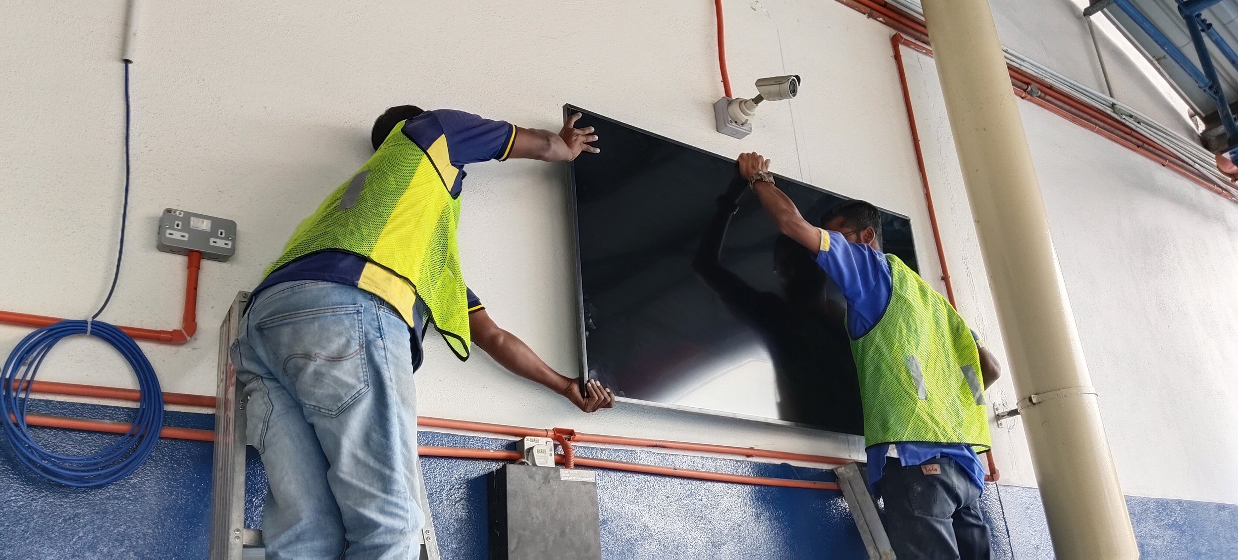 Two workers in yellow safety vests and blue shirts installing a black flat-screen TV on a white wall.