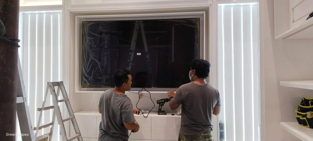 Two men working on a flat-screen TV mounted on wall in a white room, one with a mask, tools on surface below