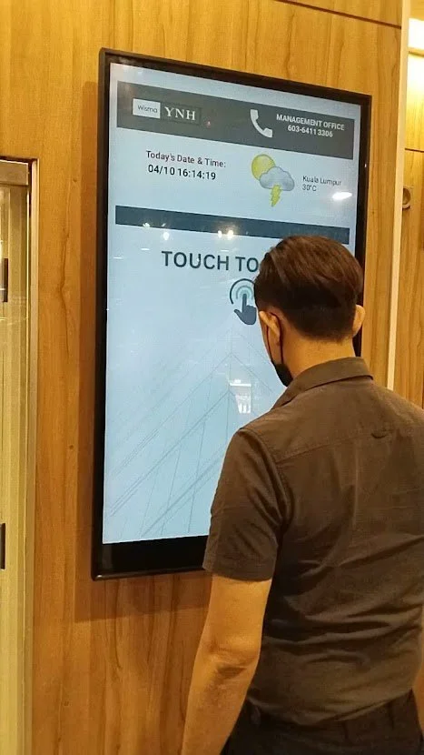 Man standing in front of a large touchscreen display in a wood-paneled room, using it with his right hand.