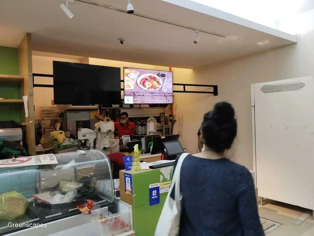 A woman with dark hair in a bun, wearing a blue sweater and carrying a white tote bag, stands in a food store. In front of her, there is a display case with various items and a green packaging box with a blue sticker. Behind the counter, a man wearin