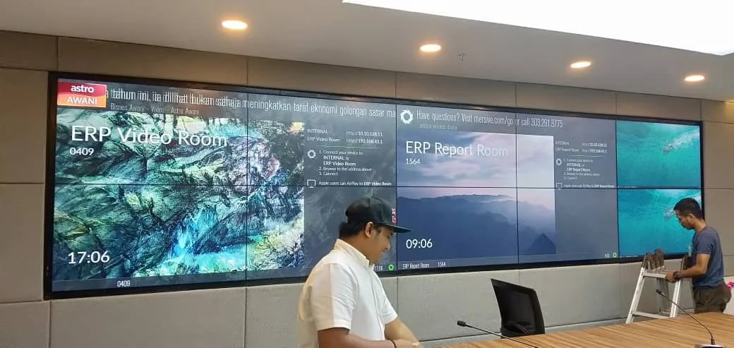 Two men are setting up or maintaining large digital display screens in a conference or control room. One man is wearing a white shirt and a cap, while the other is in a blue shirt and standing on a ladder. The screens display various information, inc