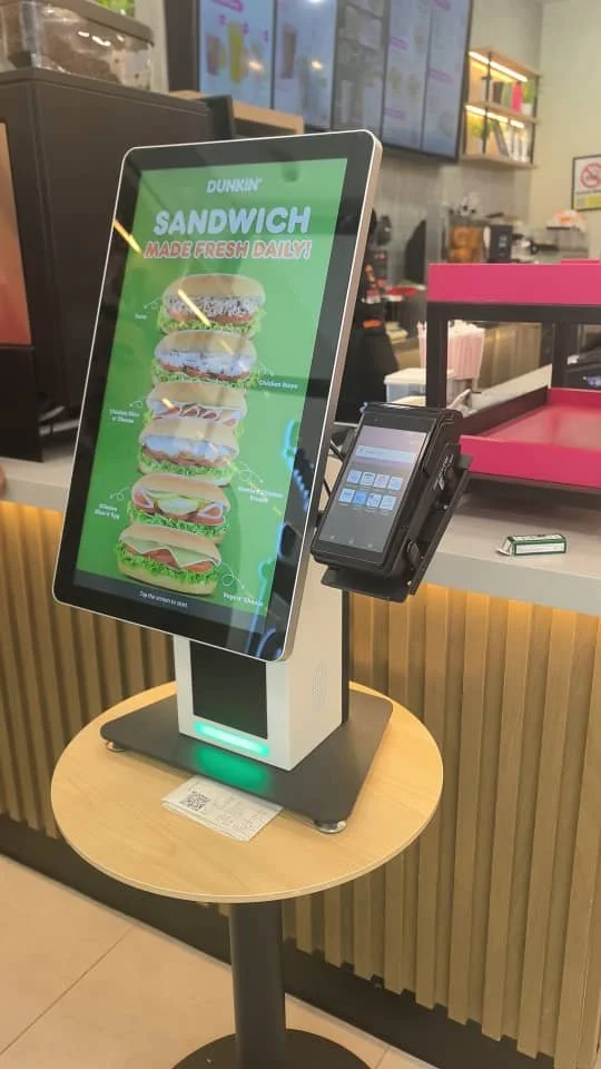Digital kiosk displaying different sandwich options at Dunkin' Donuts with a payment terminal attached