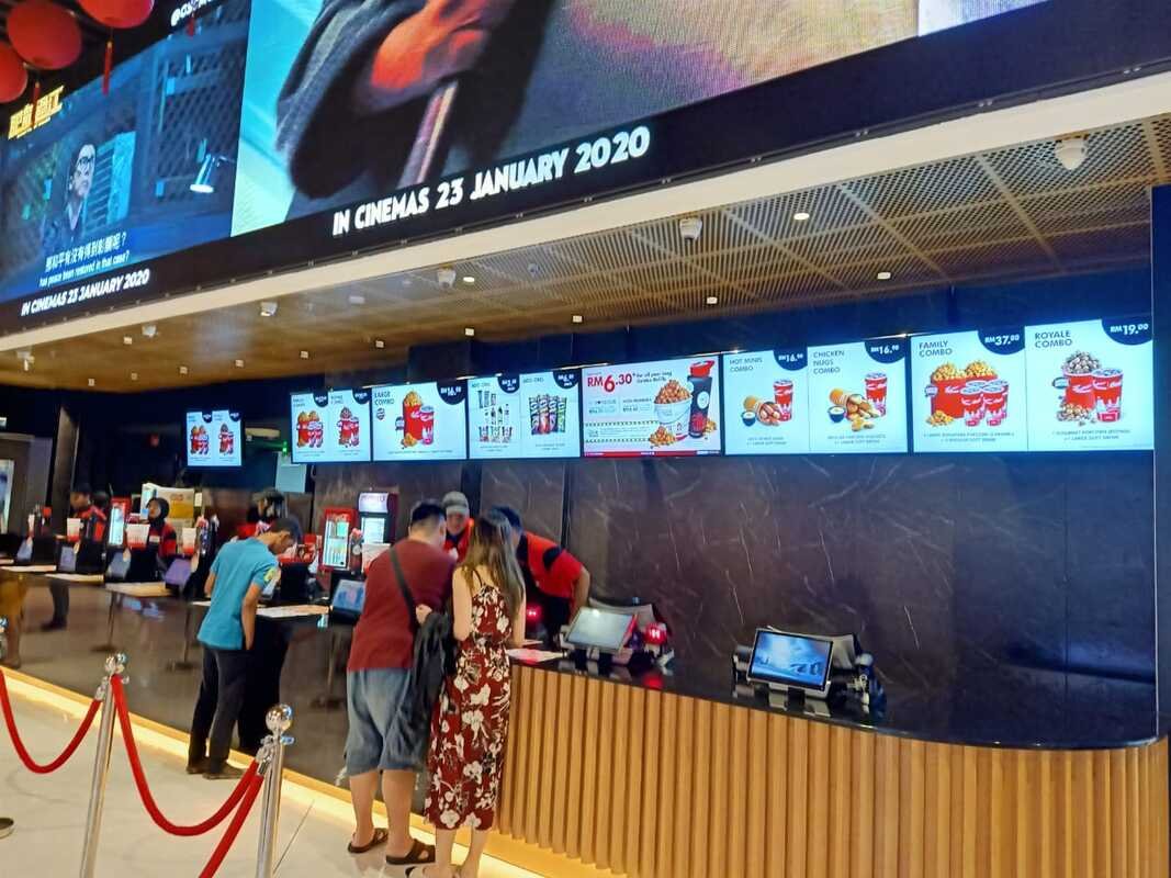 Movie theater concession stand with digital menu screens, people ordering, and a large electronic billboard displaying movie release date.