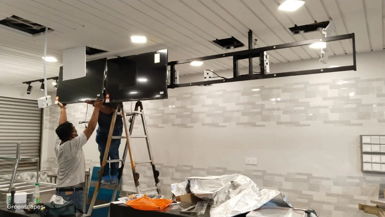 Two men installing or repairing a large flat-screen TV mounted on a ceiling mount in an indoor setting, with tools and equipment around them.