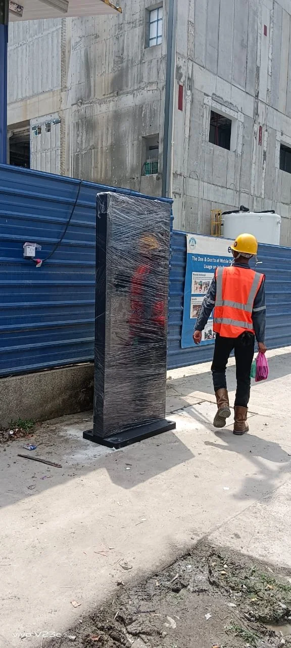Construction worker in safety vest and helmet walking past a black, wrapped, free-standing digital kiosk on a construction site sidewalk.