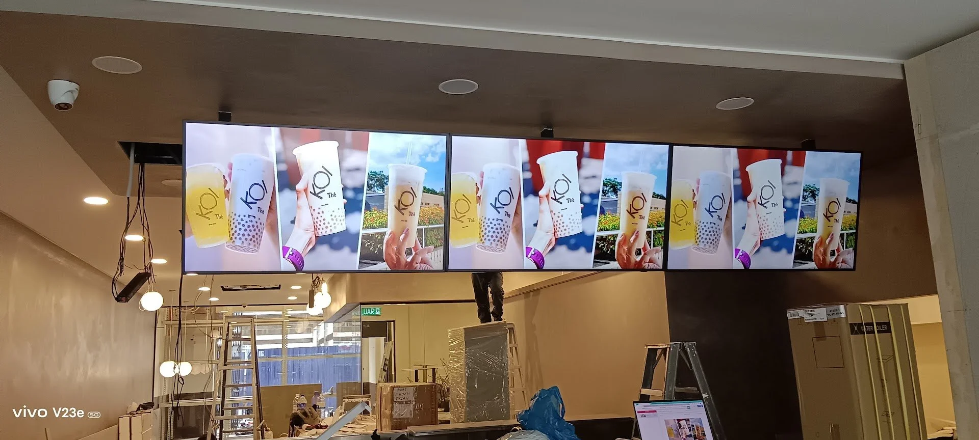 Multiple digital screens display images of hand holding cups of bubble tea with colorful backgrounds and text that reads 'KO! The'. The scene appears to be inside a store under renovation with ladders, packages, and workers around.