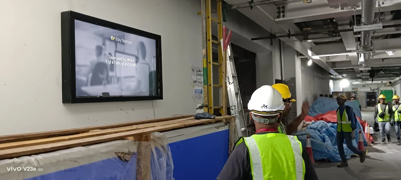 Construction workers in yellow safety vests and helmets on a construction site, with plastic-covered materials and a ladder, watching a wall-mounted digital screen displaying a downloading progress bar.