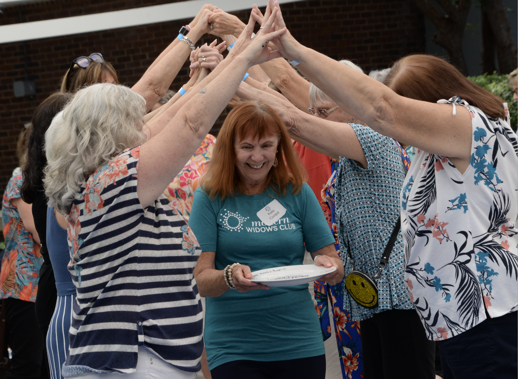 Woman wearing a turquoise Modern Widows Club t-shirt carrying a plate walks beneath a canopy created by other women with raised arms, during a MWC Release Ceremony